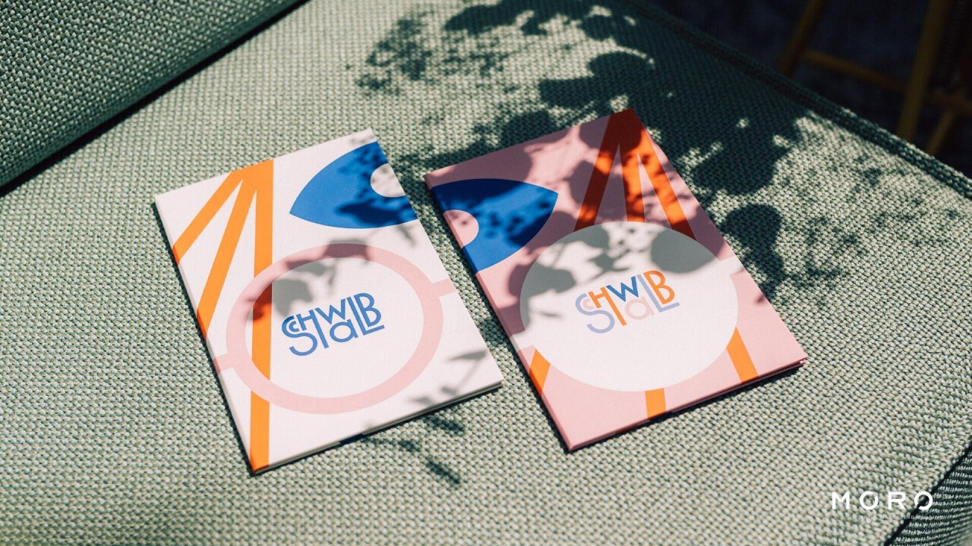 Two colorful, geometric-patterned notebooks with the text SHWB SHAB on each cover are placed side by side on a textured green fabric surface, with dappled sunlight and leaf shadows falling across them.