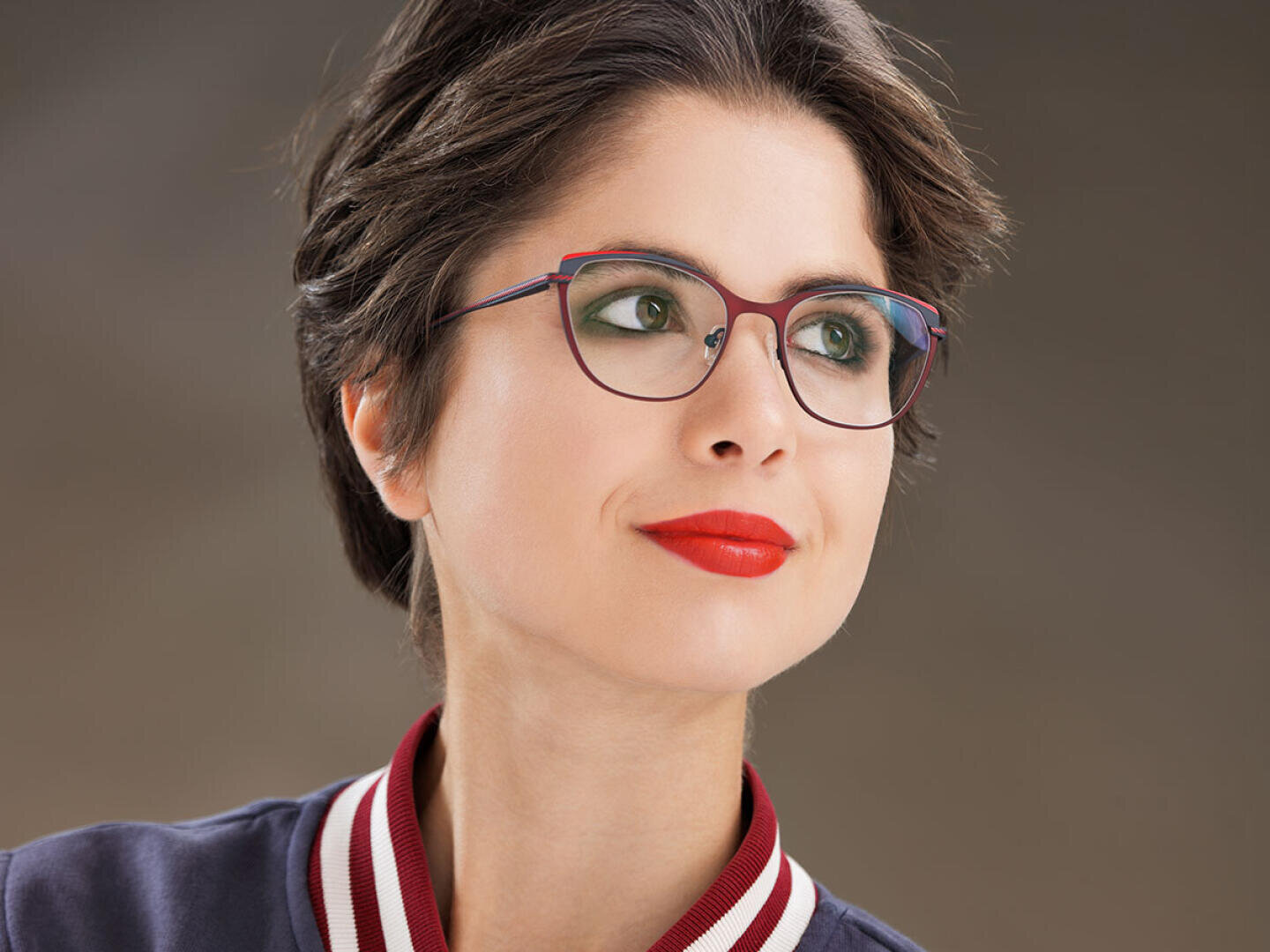 Eine Frau mit kurzen braunen Haaren, rotem Lippenstift und grünem Augen-Make-up trägt eine rot-blaue Brille. Sie hat ein leichtes Lächeln und schaut zur Seite. Sie trägt eine dunkle Jacke mit einem weiß-rot gestreiften Kragen.