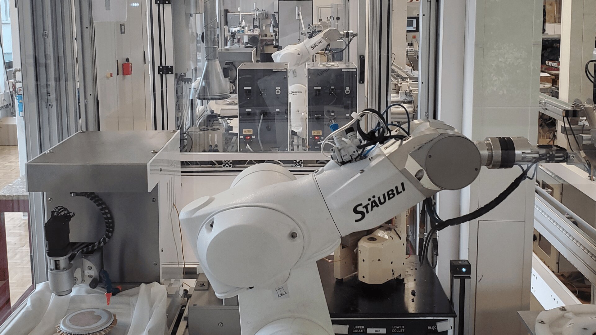 Ein weißer Staubli-Industrieroboterarm arbeitet in einem Labor oder einer Fabrik, umgeben von Glasscheiben und anderen Robotergeräten im Hintergrund.