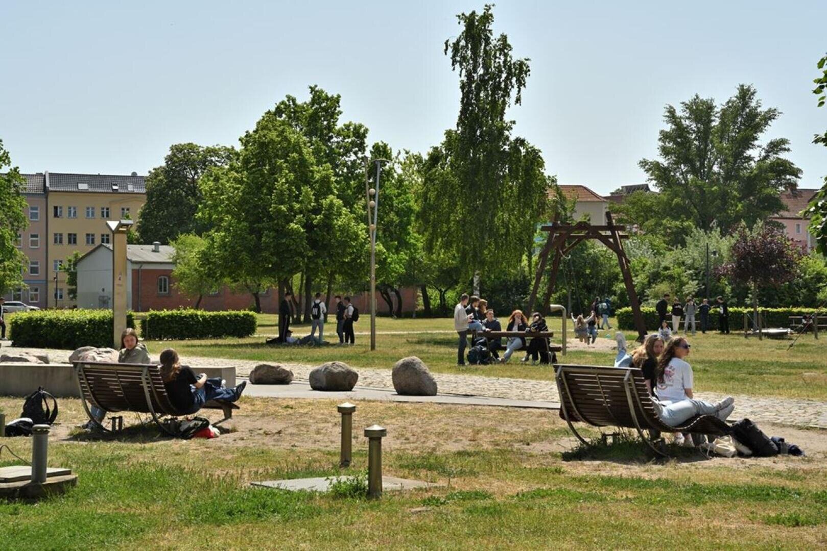 Menschen entspannen sich auf geschwungenen Bänken und Rasenflächen in einem sonnigen Park mit Bäumen, Steinen und einer Schaukel. Im Hintergrund sind Wohnhäuser und Gruppen von Menschen zu sehen, die sich unterhalten.