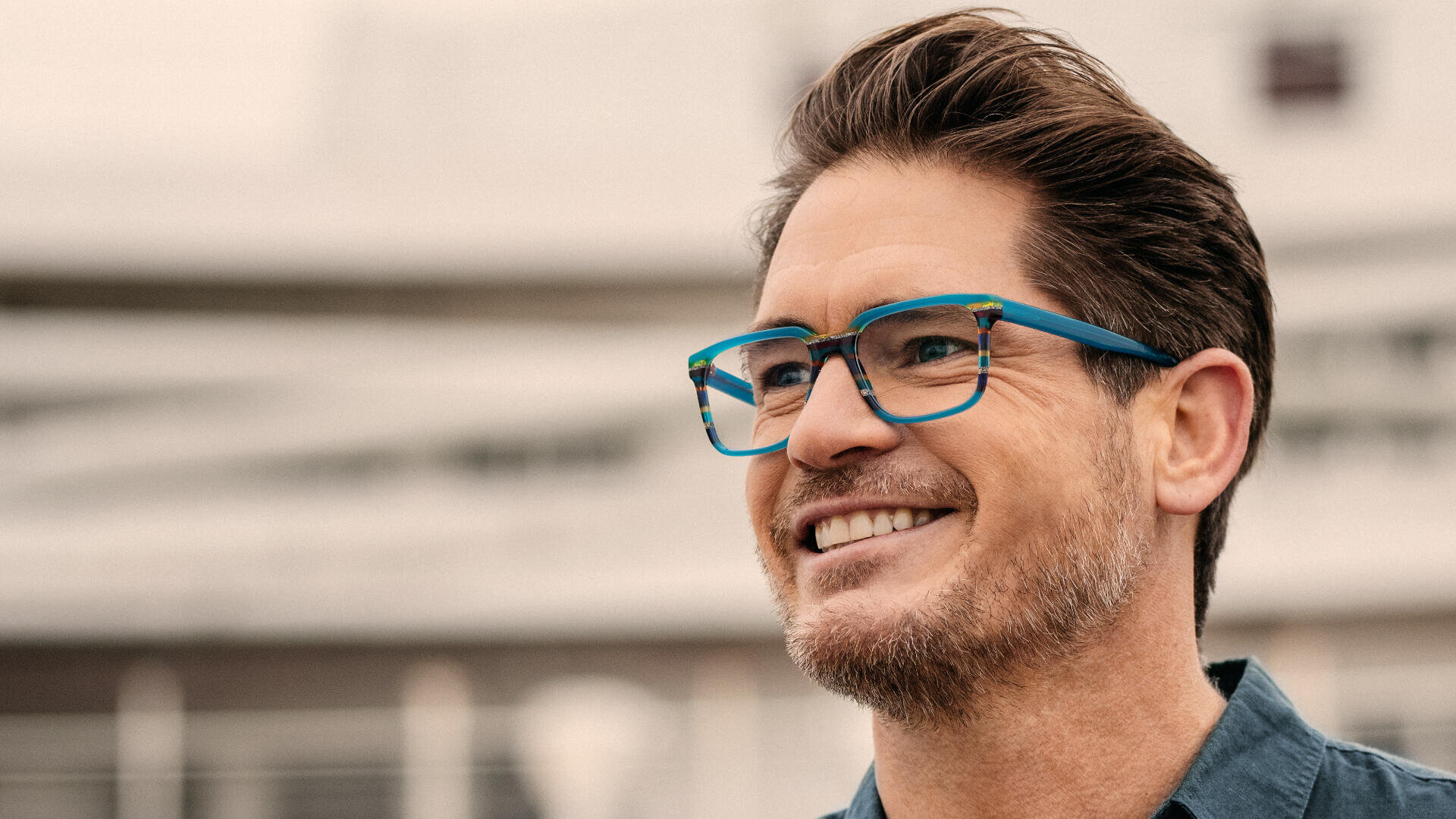 Ein lächelnder Mann mit kurzen braunen Haaren und Bart, einer Brille mit blauem Rand und einem blauen Hemd steht im Freien vor einem unscharfen Hintergrund.