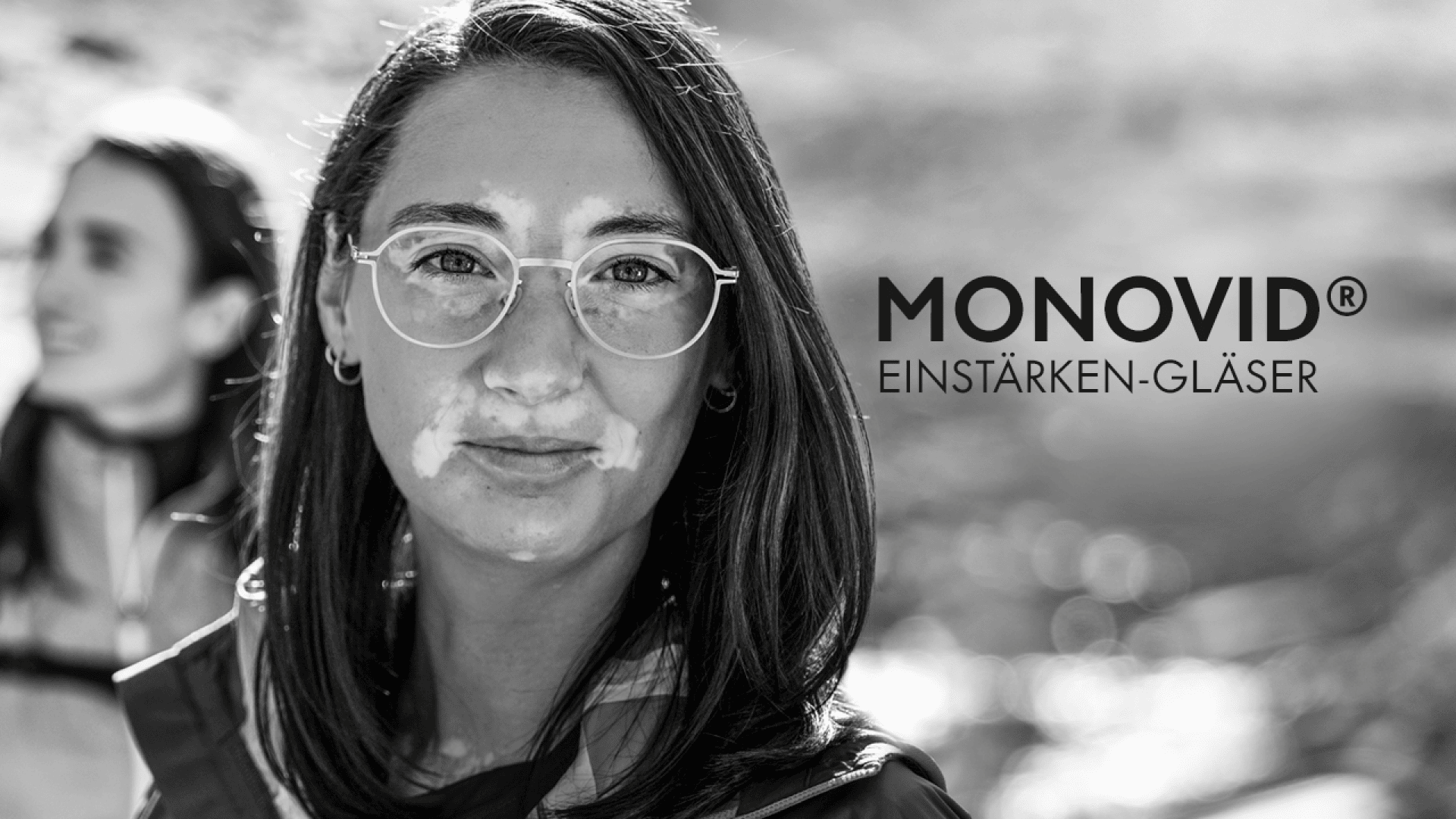 Schwarz-Weiß-Bild einer Frau mit Brille, die in die Kamera schaut, mit einer anderen Person im Hintergrund, die unscharf ist. Der Text auf der rechten Seite lautet MONOVID® Einstärken-Gläser.