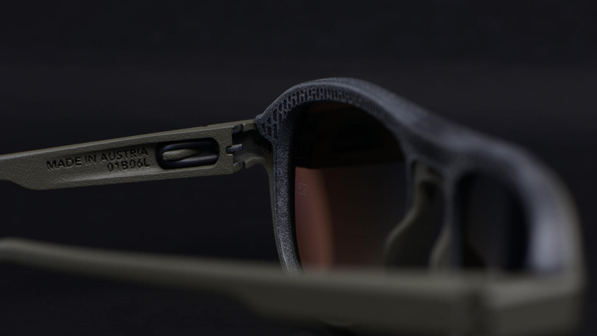 Close-up of a pair of sunglasses with dark lenses and matte green frames. The inside of the temple arm is engraved with MADE IN AUSTRIA 018106L against a black background.