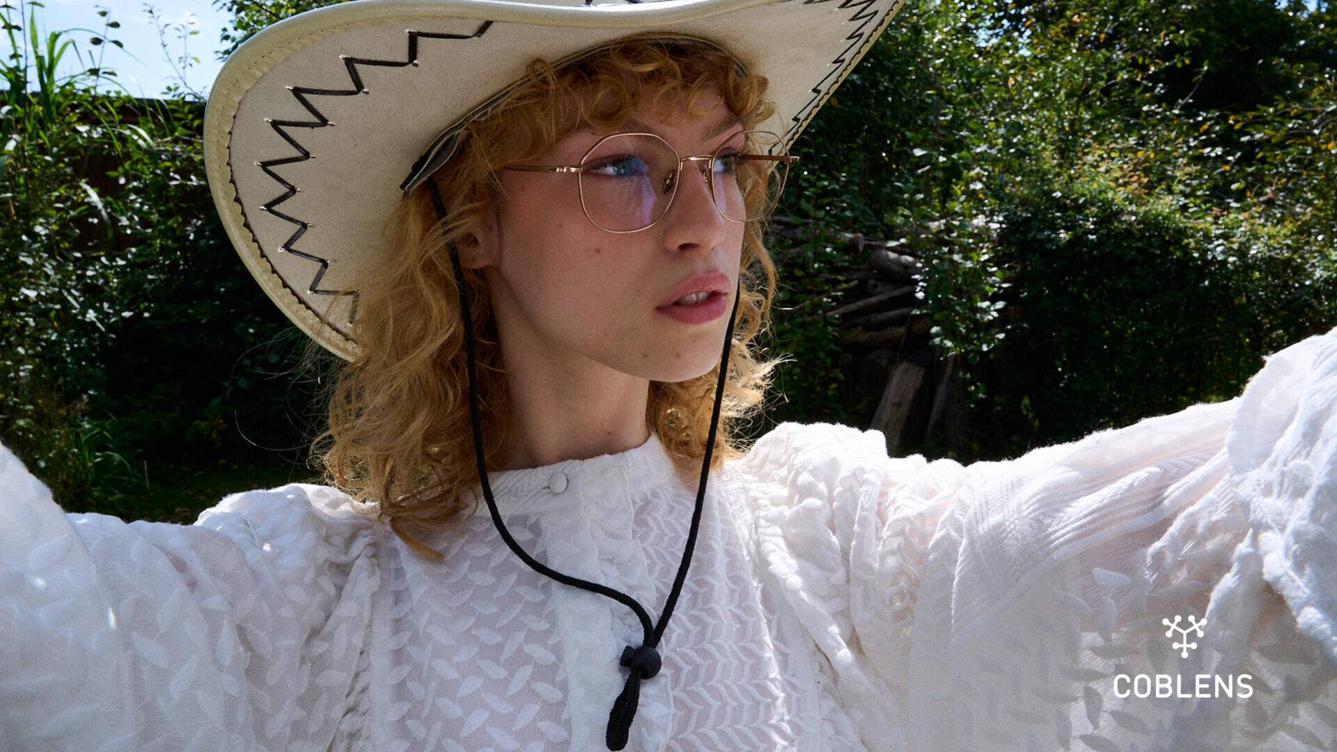A person with curly blond hair wearing glasses, a white textured blouse, and a wide-brimmed cowboy hat takes a selfie outdoors. Sunlight and greenery are visible in the background. The logo “COBLENS” appears in the corner.