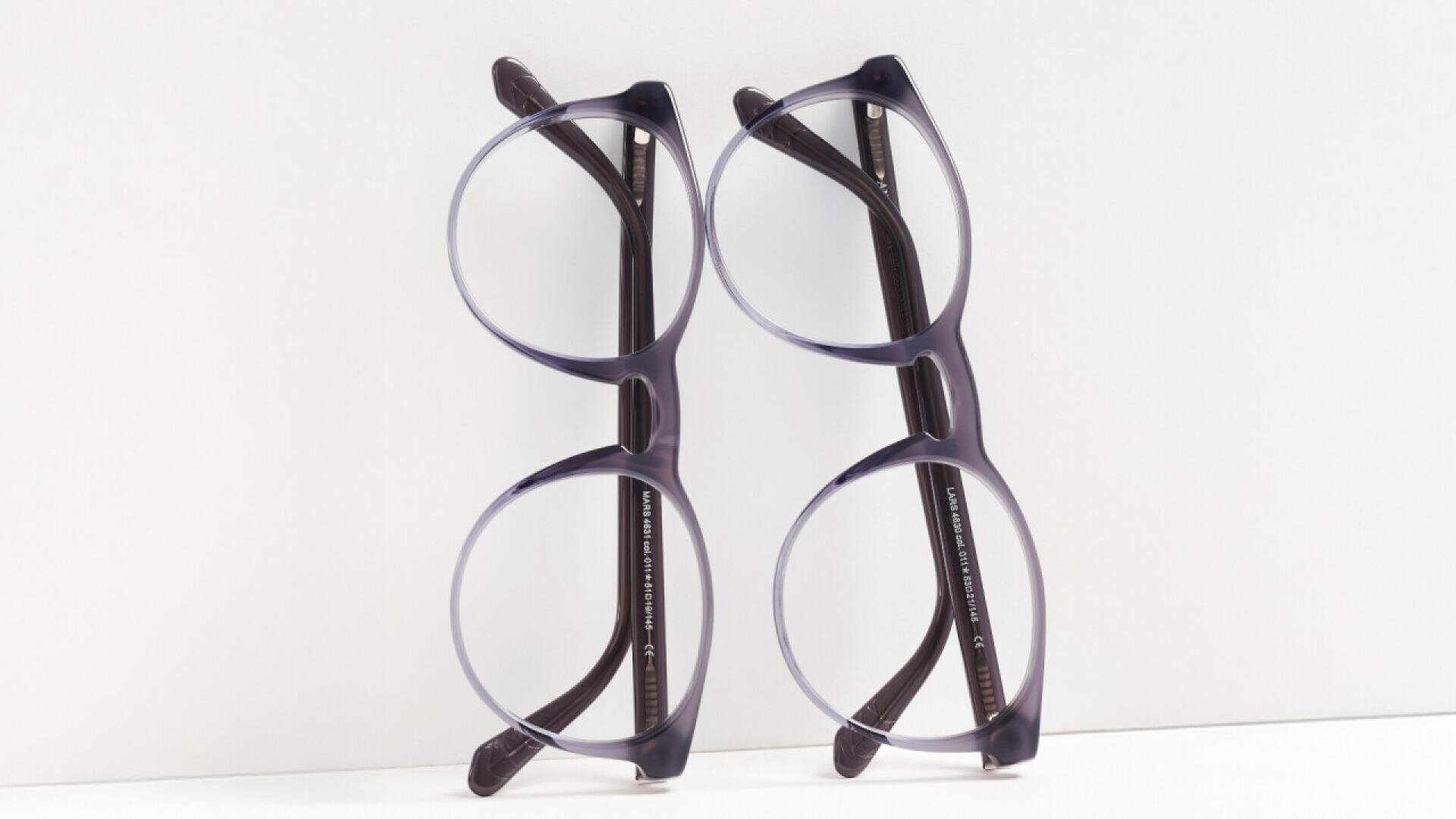 Two pairs of black-rimmed eyeglasses are placed side by side, leaning against a white background, with the temples slightly open and the glasses angled toward each other.