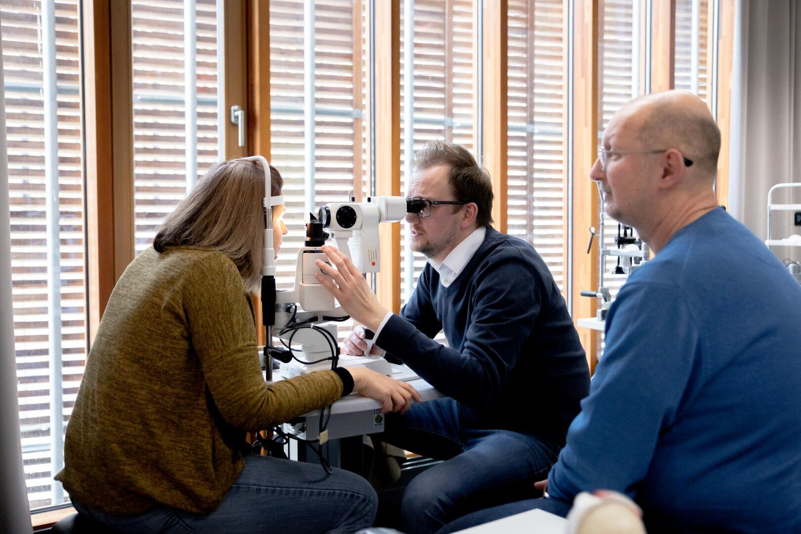 A woman is having her eyes examined with a slit lamp by a man wearing sunglasses, while another man sits nearby, in a well-lit room with large windows.