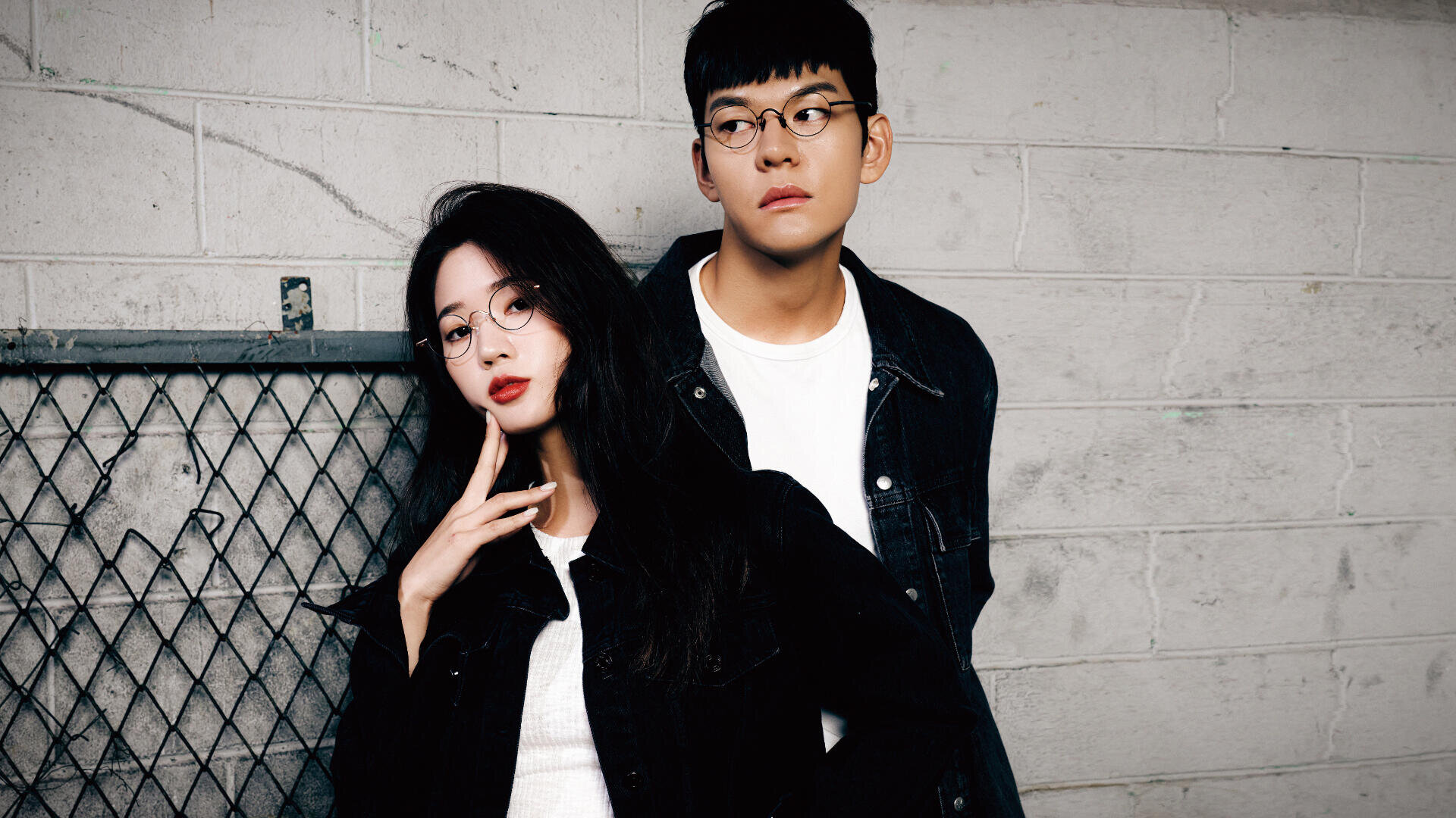 Two young adults wearing glasses and black denim jackets pose against a light gray brick wall. The woman stands in front touching her face, while the man stands slightly behind, looking to the side.