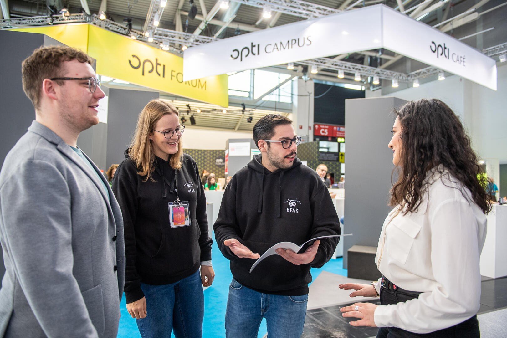 Vier Personen stehen auf einer Indoor-Messe oder -Konferenz im Gespräch. Zwei von ihnen tragen schwarze Kapuzenpullis mit einem Logo, einer hält ein aufgeschlagenes Heft in der Hand, ein anderer trägt ein Abzeichen. Im Hintergrund sind Messestände und Schilder mit der Aufschrift "opti" zu sehen.