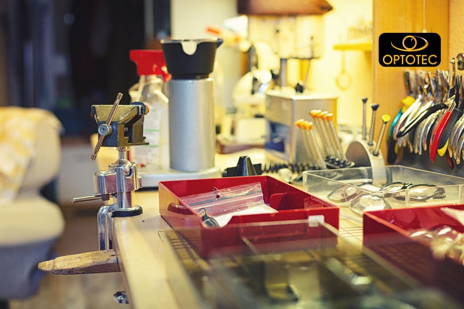 Eine Nahaufnahme einer vollgestopften Werkbank mit Werkzeugen, Ablagen und Gläsern in einer gut beleuchteten Werkstatt. Das OPTOTEC-Logo erscheint in der oberen rechten Ecke.