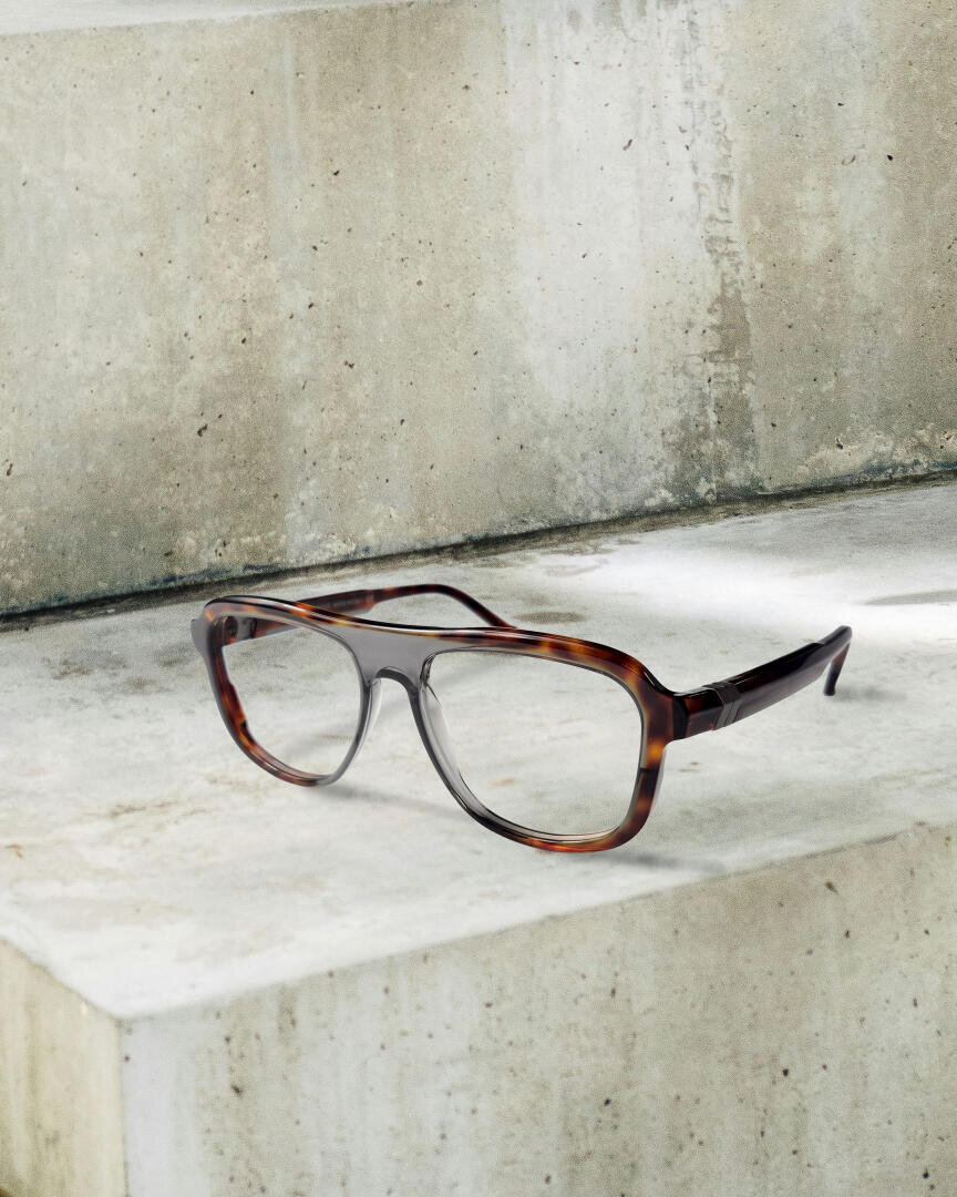 A pair of tortoiseshell glasses with a large, rectangular frame rests on a concrete surface with rough, light gray structures in the background.
