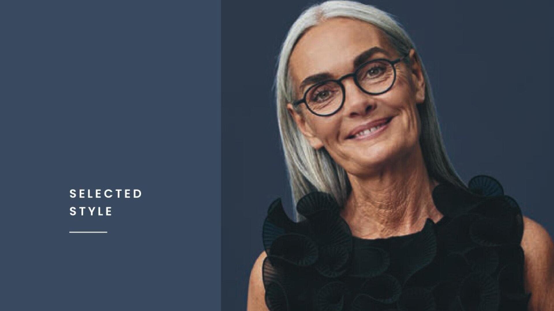 Elegant older woman with long gray hair and glasses, smiling gently, wearing a black textured top against a dark blue background. The words SELECTED STYLE appear on the left side.