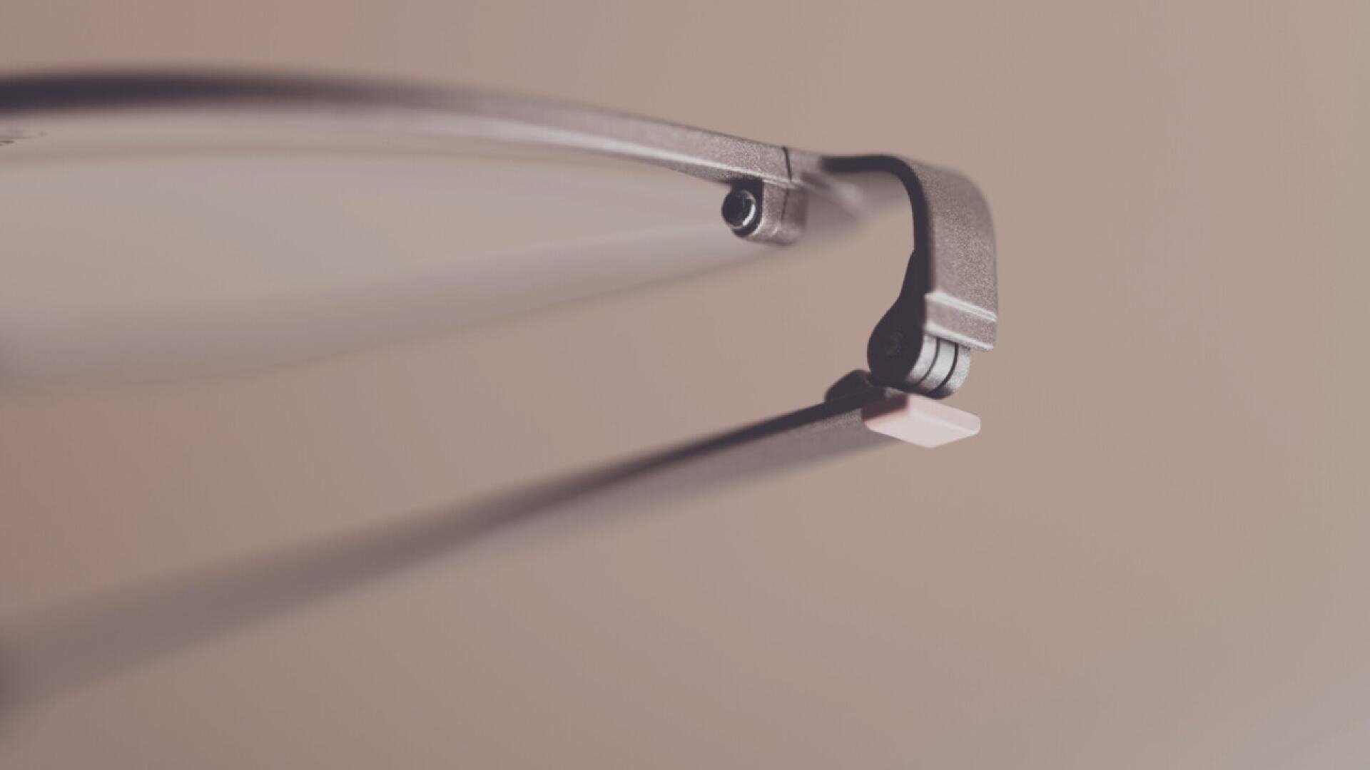 Close-up of the hinge and temple arm of a pair of eyeglasses, shot with a shallow depth of field against a muted background.