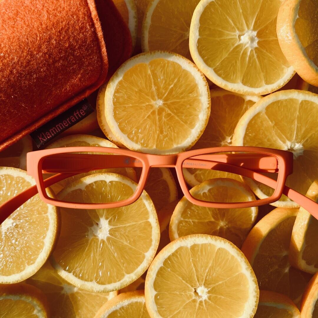 Eine Brille mit orangefarbenem Rahmen liegt auf mehreren übereinanderliegenden Zitronenscheiben, wobei in der oberen linken Ecke teilweise ein orangefarbenes Brillenetui sichtbar ist.