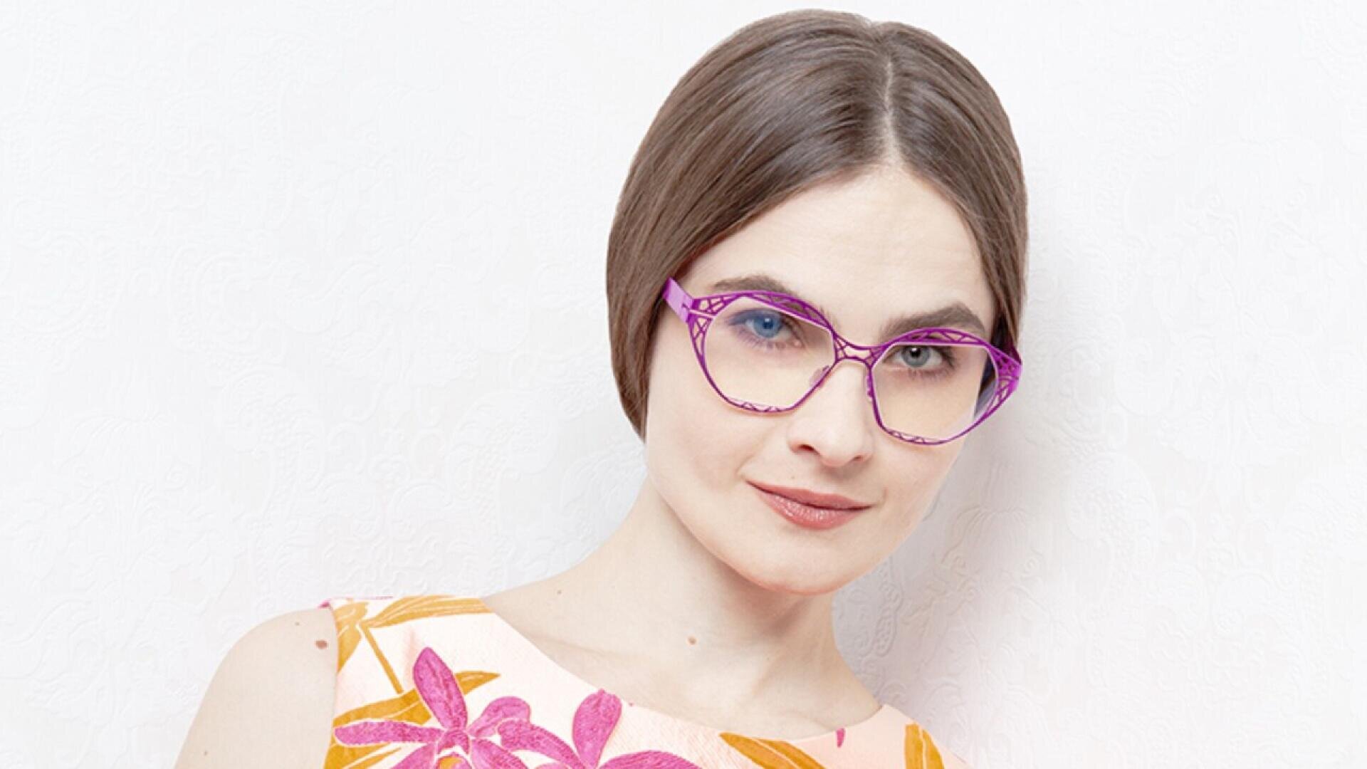 A woman with short brown hair wearing bright pink eyeglasses and a sleeveless top with an orange and pink floral pattern, posing against a white textured background.