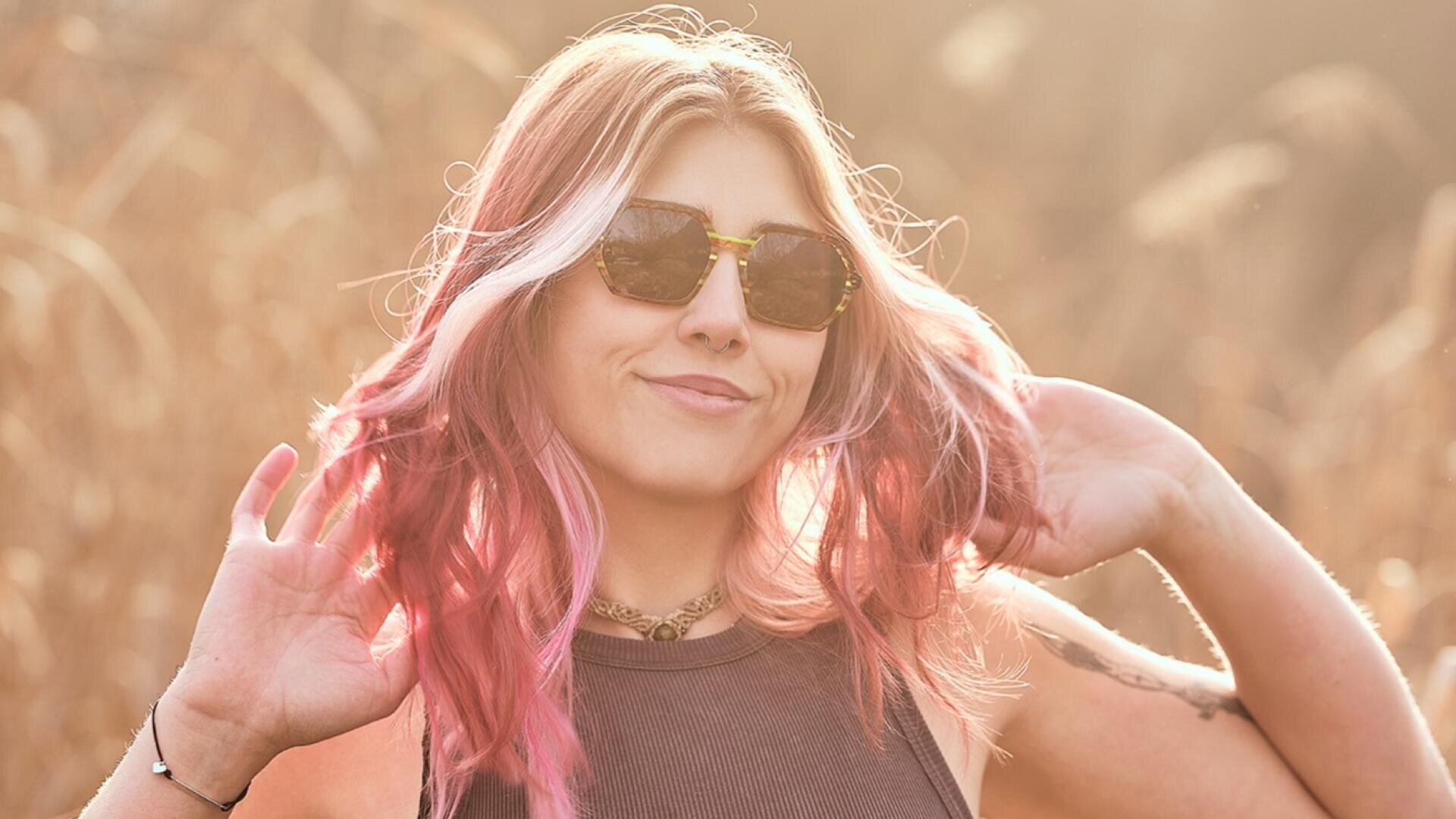 Eine Frau mit gewelltem rosafarbenem Haar und Sonnenbrille lächelt im Freien im Sonnenlicht, sie trägt ein ärmelloses Oberteil und eine Halskette, eine Hand in ihrem Haar und einen unscharfen grasbewachsenen Hintergrund.
