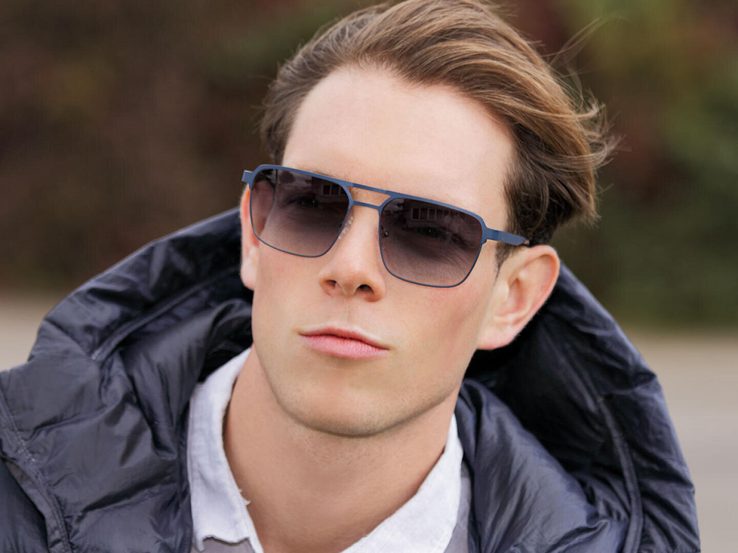 A young man with fair skin and styled brown hair wears sunglasses, a white collared shirt and a dark puffer jacket. He looks confidently into the camera with a neutral expression. The background is blurred.