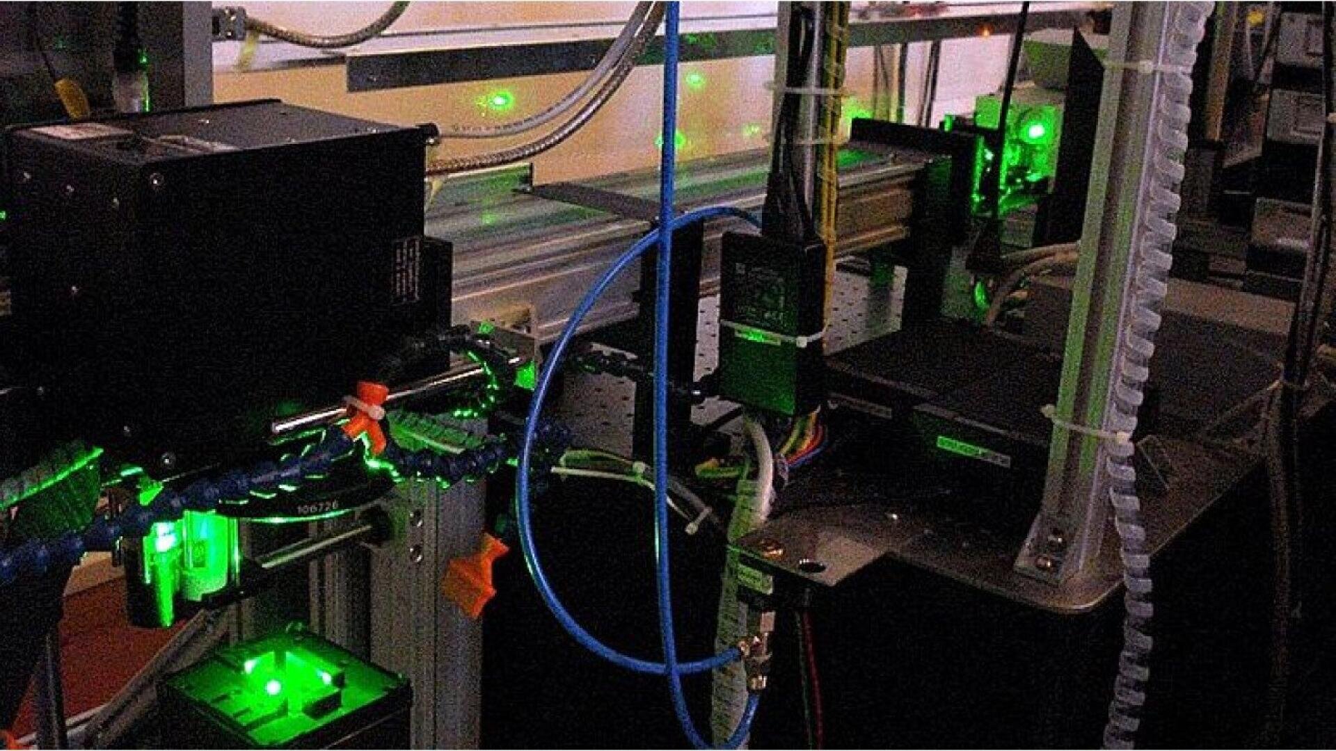 A close-up of laboratory equipment featuring various cables, mechanical components, and devices with visible green laser lights, used for scientific or technological experiments.