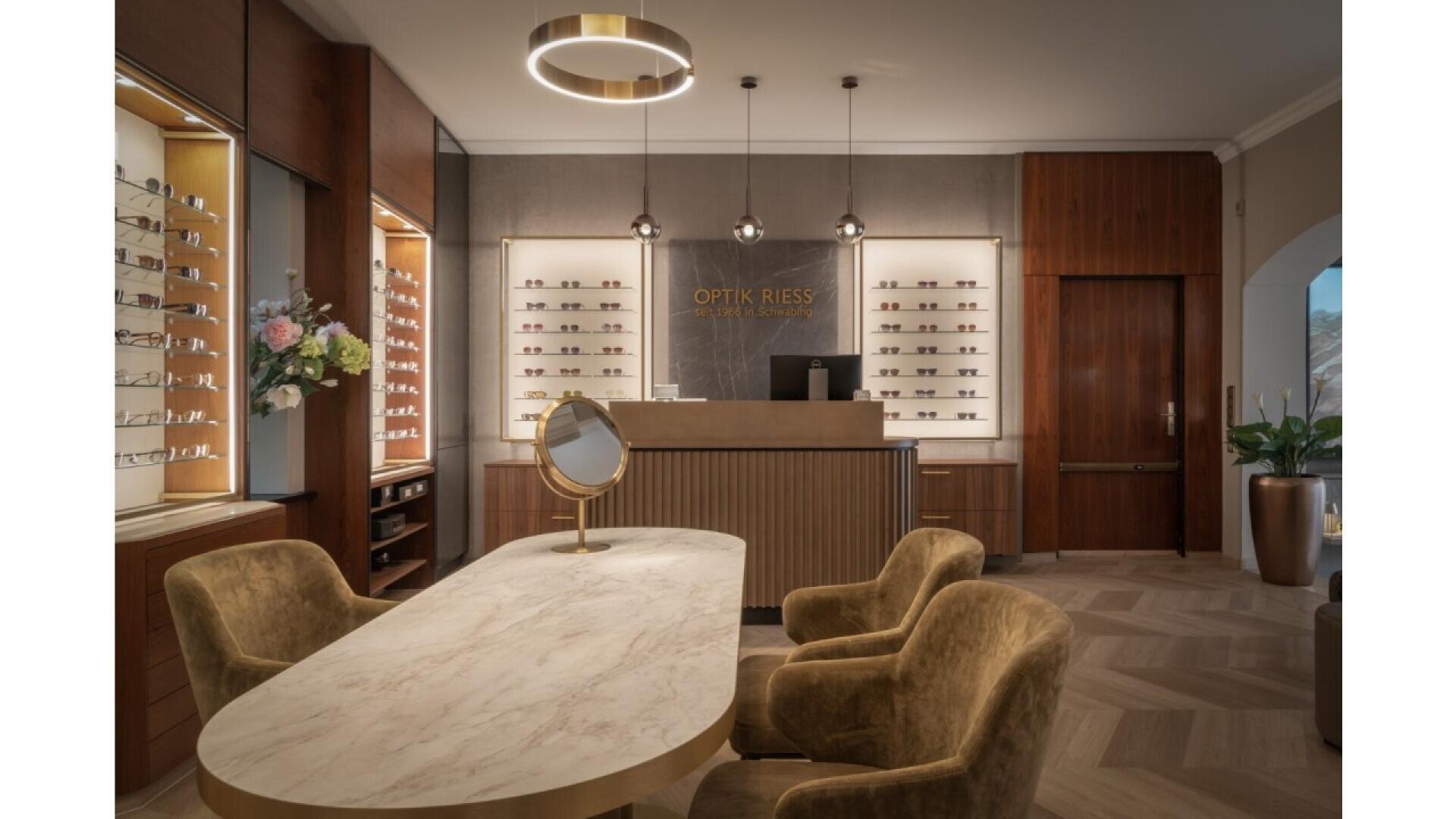 A stylish opticians office with a marble table, velvet chairs, wall displays of eyeglasses, flower arrangement, a gold-framed mirror, and modern pendant lighting above a wooden reception counter.