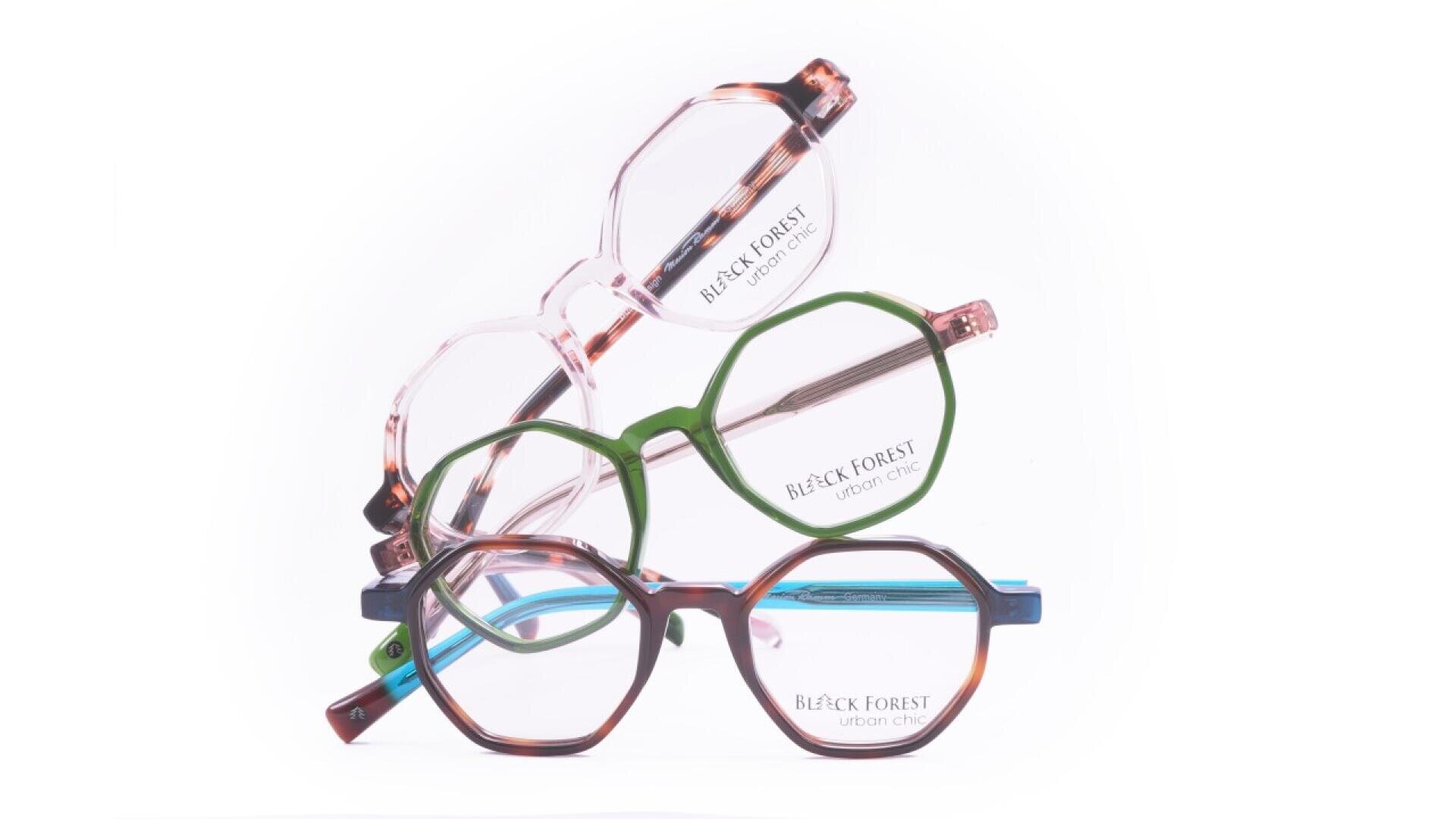Three pairs of eyeglasses with round and geometric frames in green, pink, and multicolor patterns are stacked on each other. The frames have BLACK FOREST URBAN OPTIC printed on the lenses. The background is white.