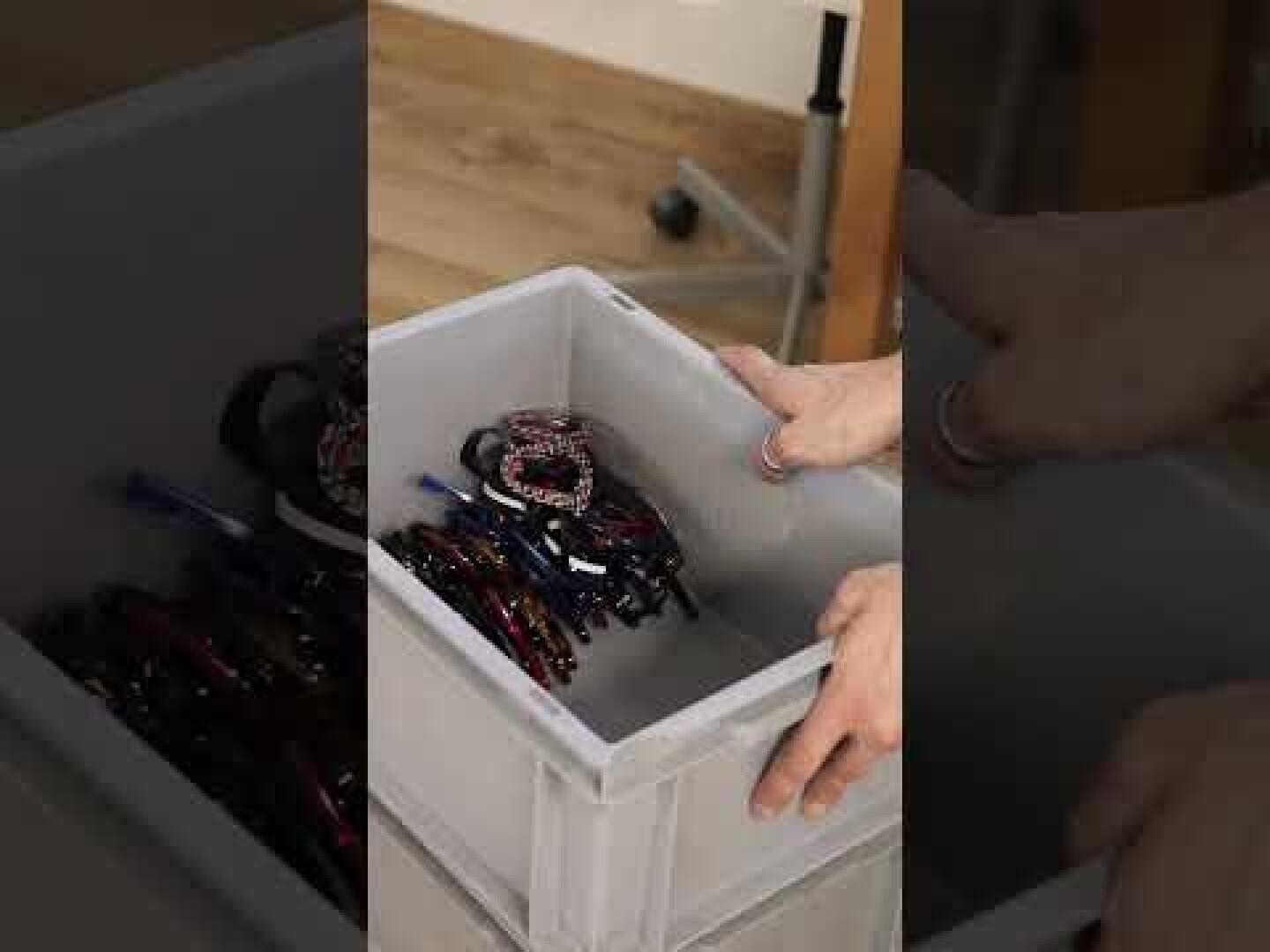 A person places a plastic container filled with assorted headbands and hair accessories into a larger gray storage bin on a wooden floor.