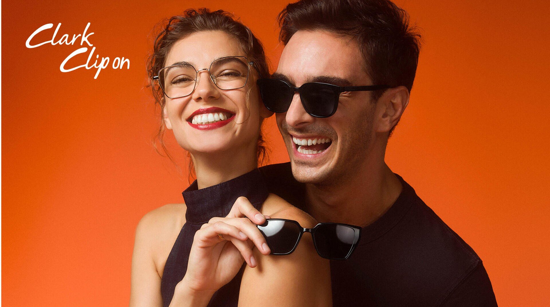 Eine lächelnde Frau mit Brille und ein Mann mit Sonnenbrille stehen dicht nebeneinander vor einem orangefarbenen Hintergrund. Die Frau hält eine weitere Sonnenbrille, und beide sehen glücklich aus. Clark Clip on steht in weißer Schrift in der Ecke.