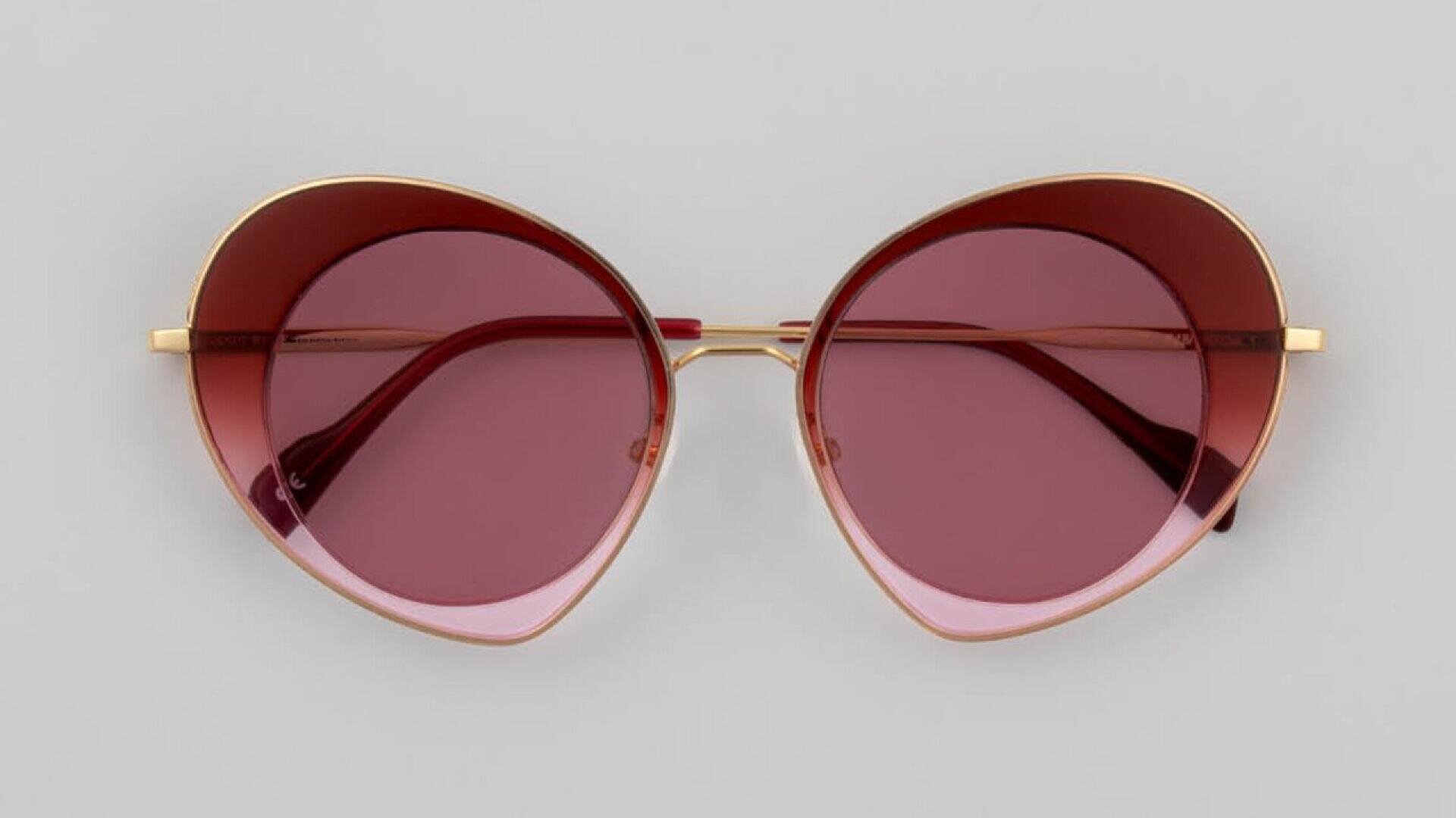 A pair of cat-eye sunglasses with gold frames and rose-tinted lenses, displayed on a light gray background.