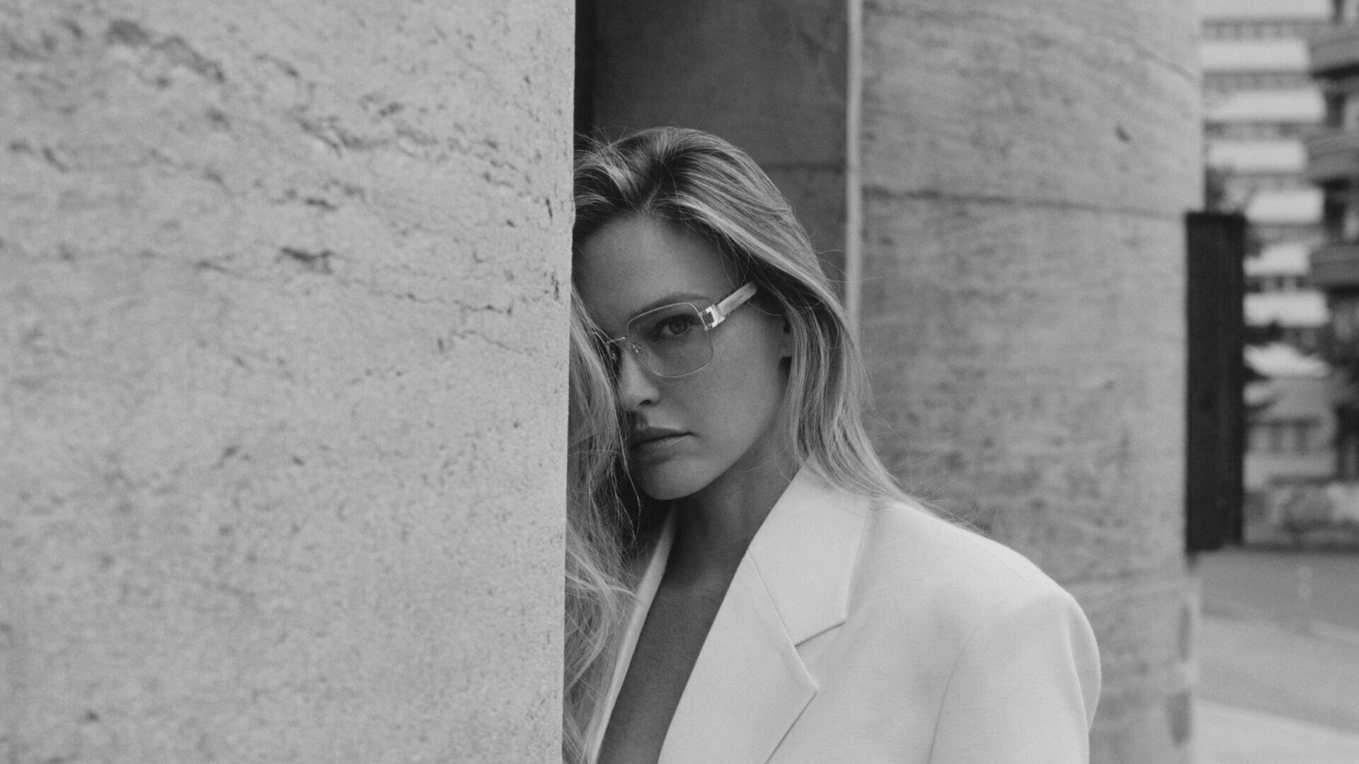 A woman in a light-colored blazer and glasses stands next to a large, textured stone column and looks into the camera. The image is in black and white with a blurred urban background.