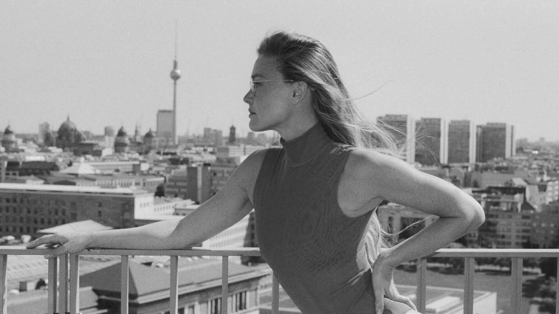 A woman with long hair and sunglasses leans against a railing and looks out over a cityscape with the Berlin television tower in the background. The photo is in black and white.