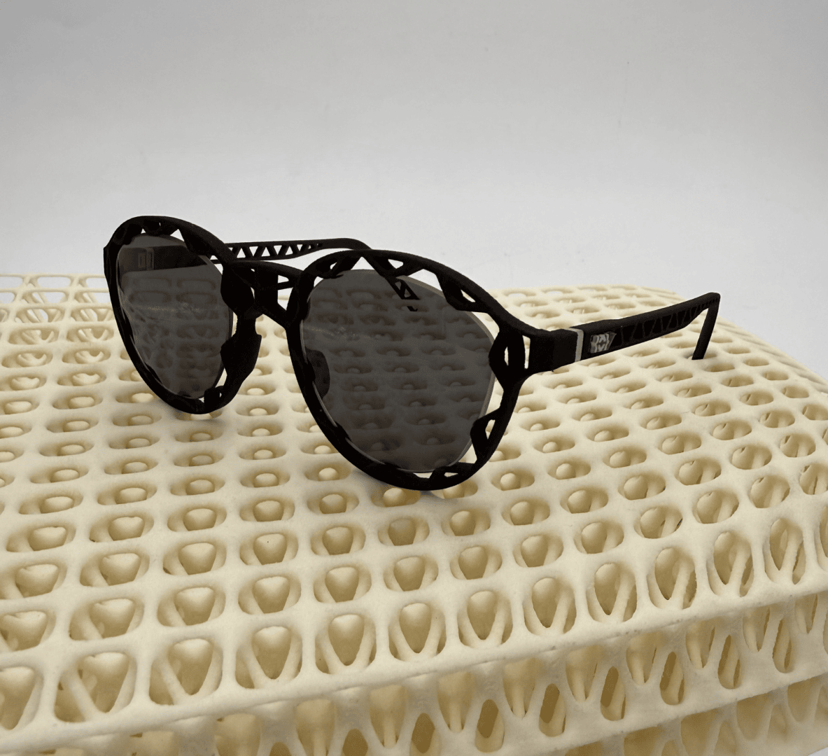 Eine runde schwarze Sonnenbrille mit einem dekorativen, gewellten Rahmen ruht auf einer cremefarbenen, gitterartig gemusterten Oberfläche vor einem einfarbig weißen Hintergrund.