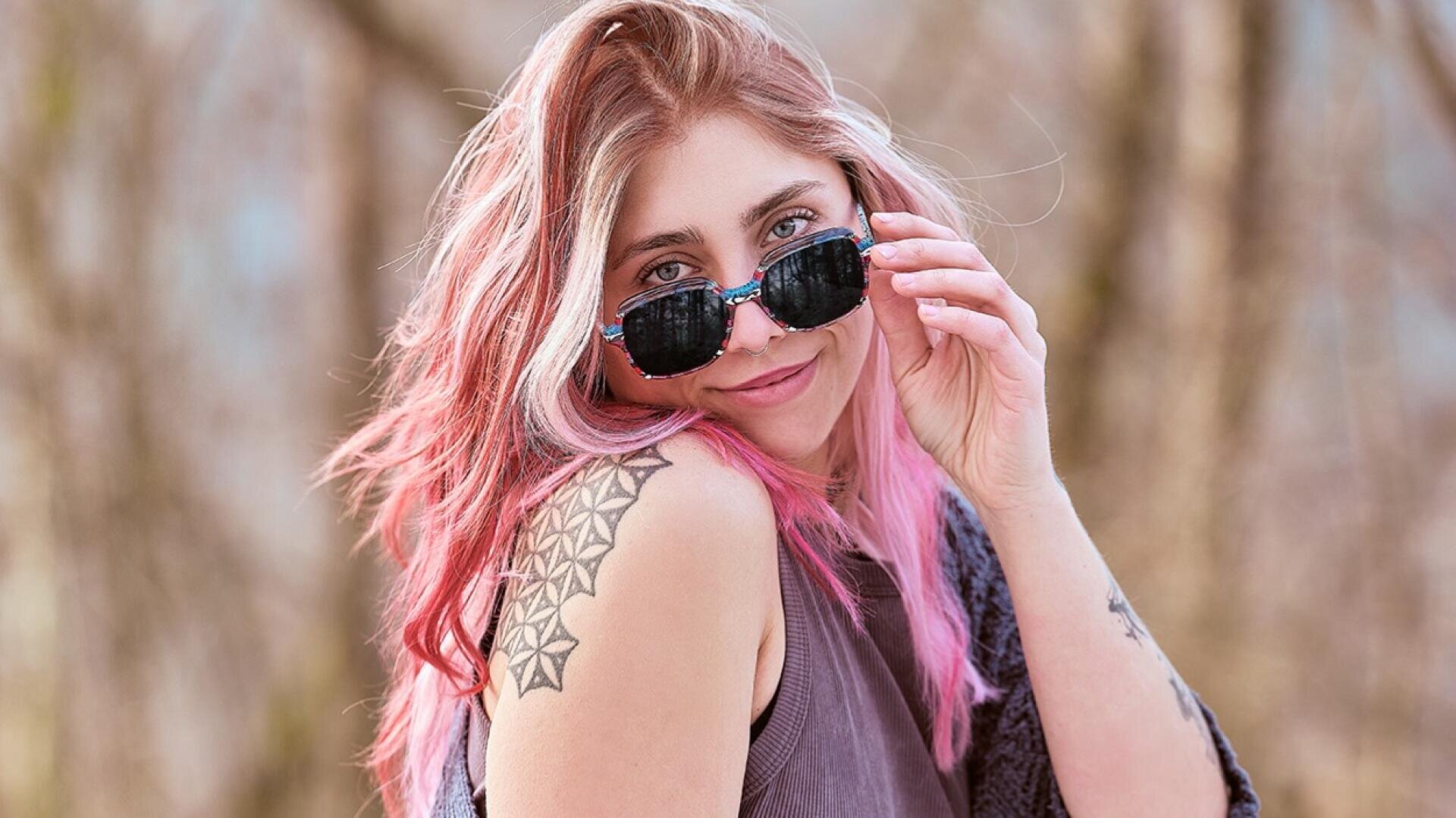 Eine junge Frau mit langen rosa Haaren und einer geometrischen Tätowierung auf der Schulter lächelt, während sie ihre Sonnenbrille hält. Sie trägt ein ärmelloses Oberteil und steht im Freien vor einem unscharfen natürlichen Hintergrund.