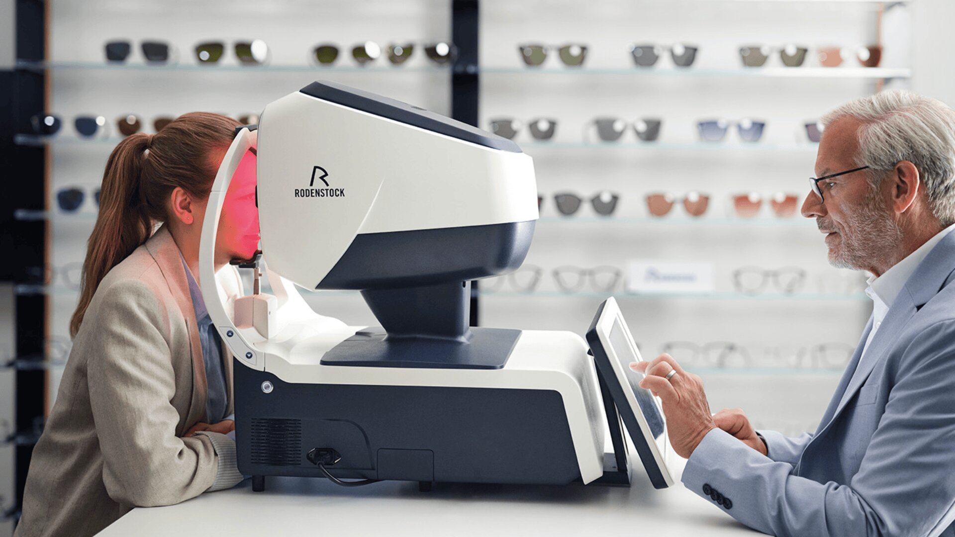 Eine Frau lässt ihre Augen von einem Augenarzt mit einem modernen Gerät in einem Optikergeschäft untersuchen, während im Hintergrund Brillenregale zu sehen sind.