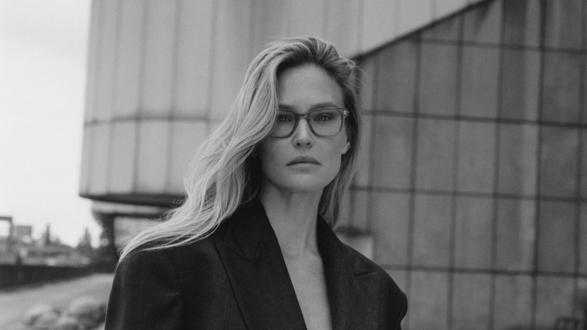 A woman with long, light-colored hair, glasses and a dark blazer stands outside in front of a modern, geometric building. The photo is black and white, and she looks directly into the camera with a calm gaze.