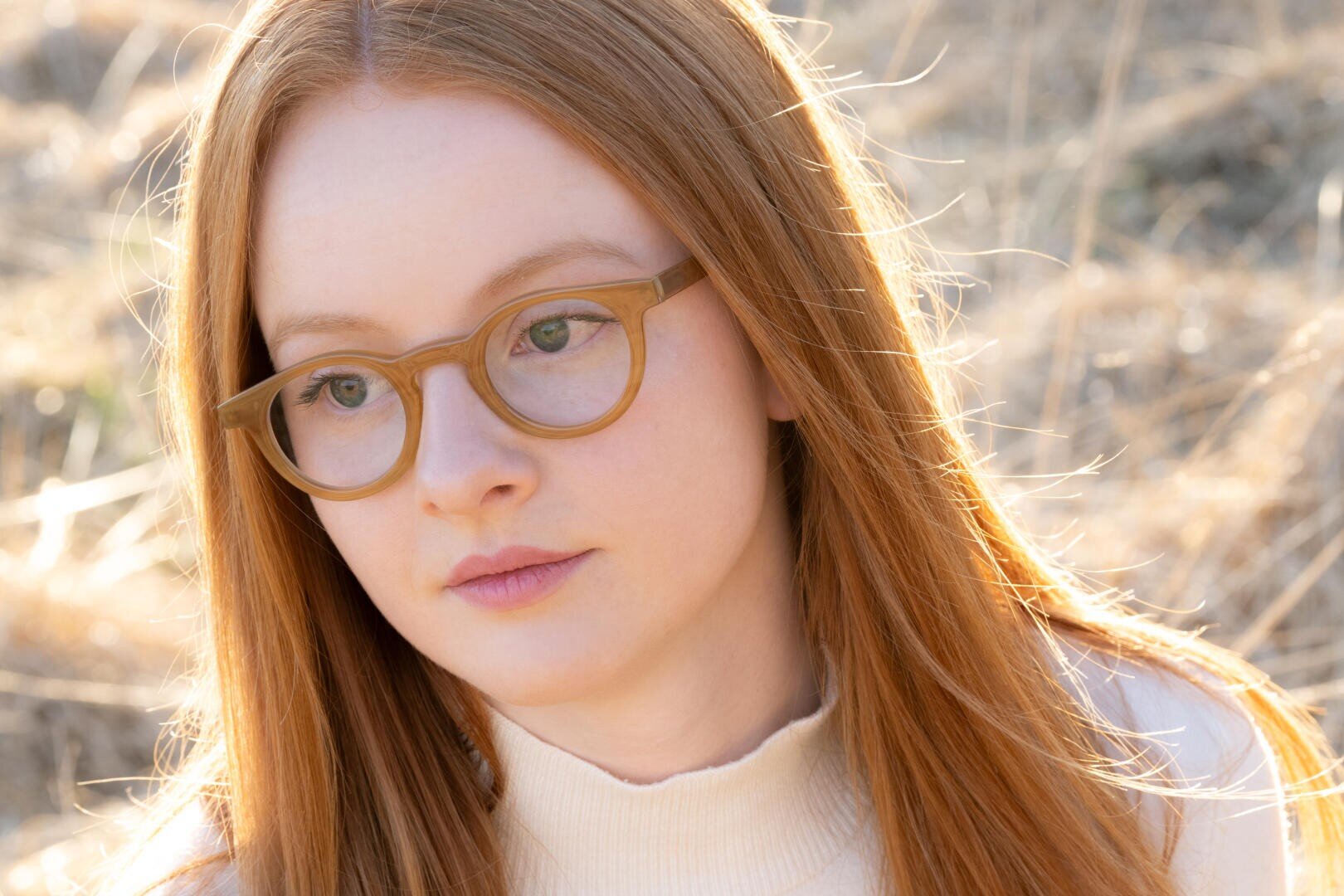 Eine junge Person mit langen roten Haaren und einer hellbraunen Brille blickt nachdenklich zur Seite ins Freie, das in warmes Sonnenlicht getaucht ist, mit trockenem Gras im Hintergrund.