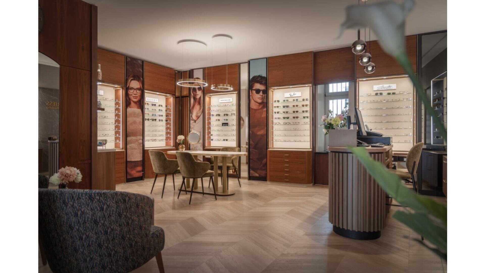 A modern optician’s store with stylish wooden decor, display shelves of eyeglasses, posters of people wearing glasses, a consultation table with chairs, and a cozy seating area in the foreground.