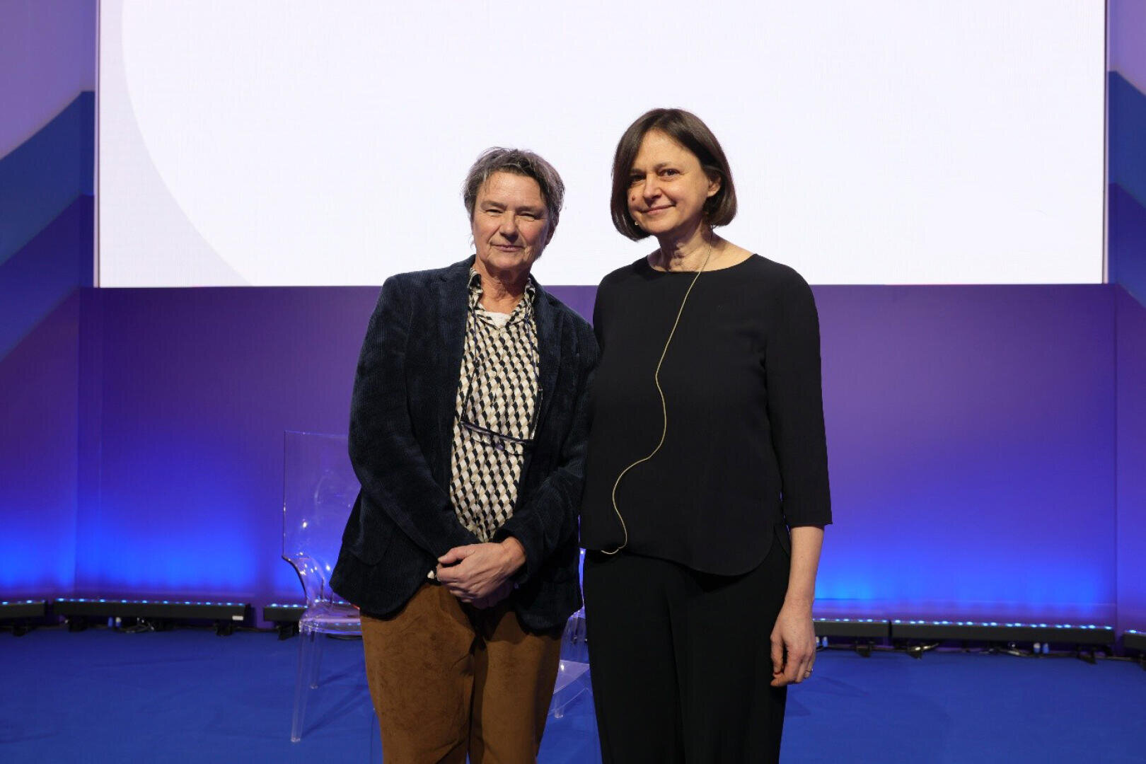 Angela Mrositzki and Francesca Valan stand next to each other on a stage with a blue and purple background. Both are looking into the camera and smiling. The person on the right is wearing a headset microphone.