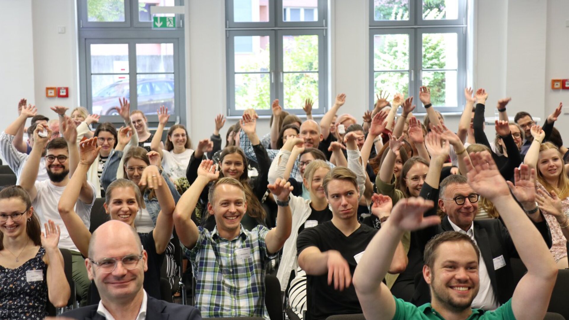 Eine große Gruppe von Menschen sitzt in einem hellen Raum, lächelt und hebt enthusiastisch die Hände, während sie in die Kamera schauen. Die Gruppe scheint auf einer Veranstaltung oder einem Seminar zu sein, im Hintergrund sind Fenster und Grünflächen zu sehen.
