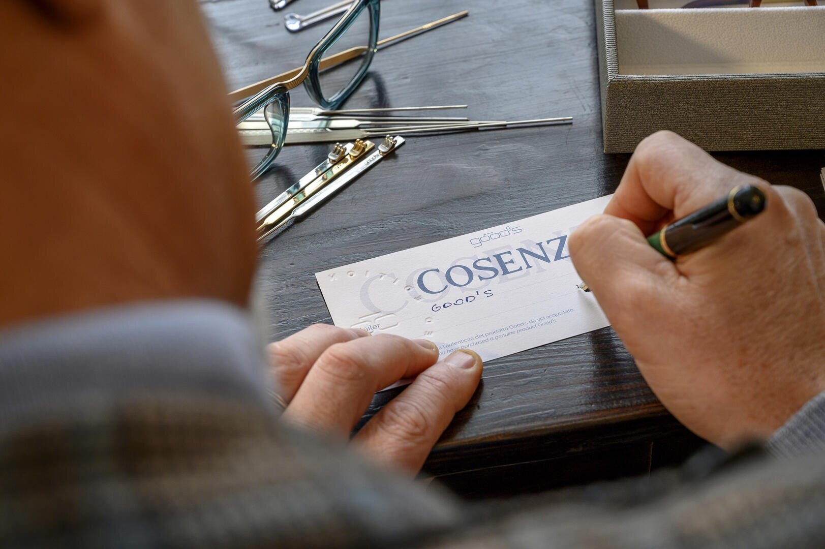 Eine Person in einer karierten Jacke schreibt an einem Schreibtisch auf eine Karte mit der Aufschrift COSENZA. In der Nähe sind Brillen, Werkzeuge und eine offene Schachtel zu sehen.