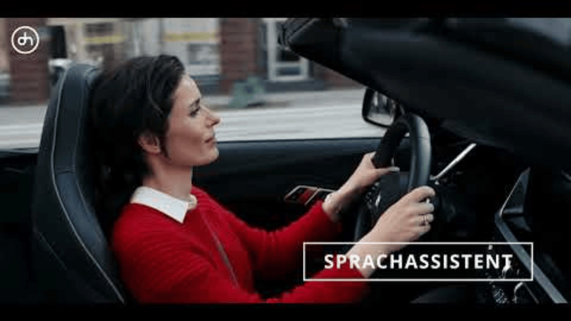 Eine Frau im roten Pullover fährt ein Cabrio mit offenem Verdeck. Sie lächelt und blickt nach vorn. Auf dem Bild erscheint das Wort „Sprachassistent“.