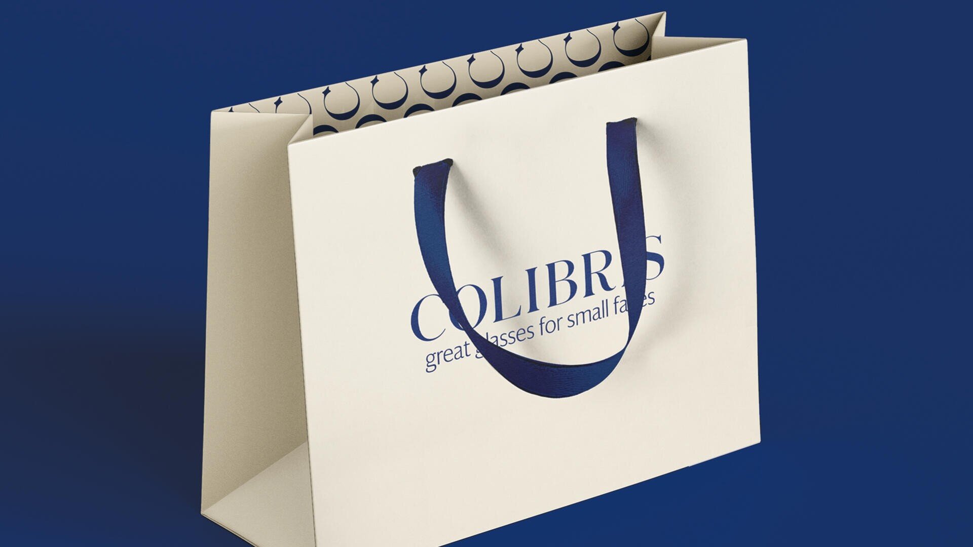 Eine cremefarbene Einkaufstasche mit blauen Henkeln. Auf der Vorderseite ist der Text „COLIBRIS – tolle Brille für kleine Gesichter“ zu sehen. Das Innenfutter zeigt ein Muster aus dunkelblauen Halbkreisen. Der Hintergrund ist marineblau.