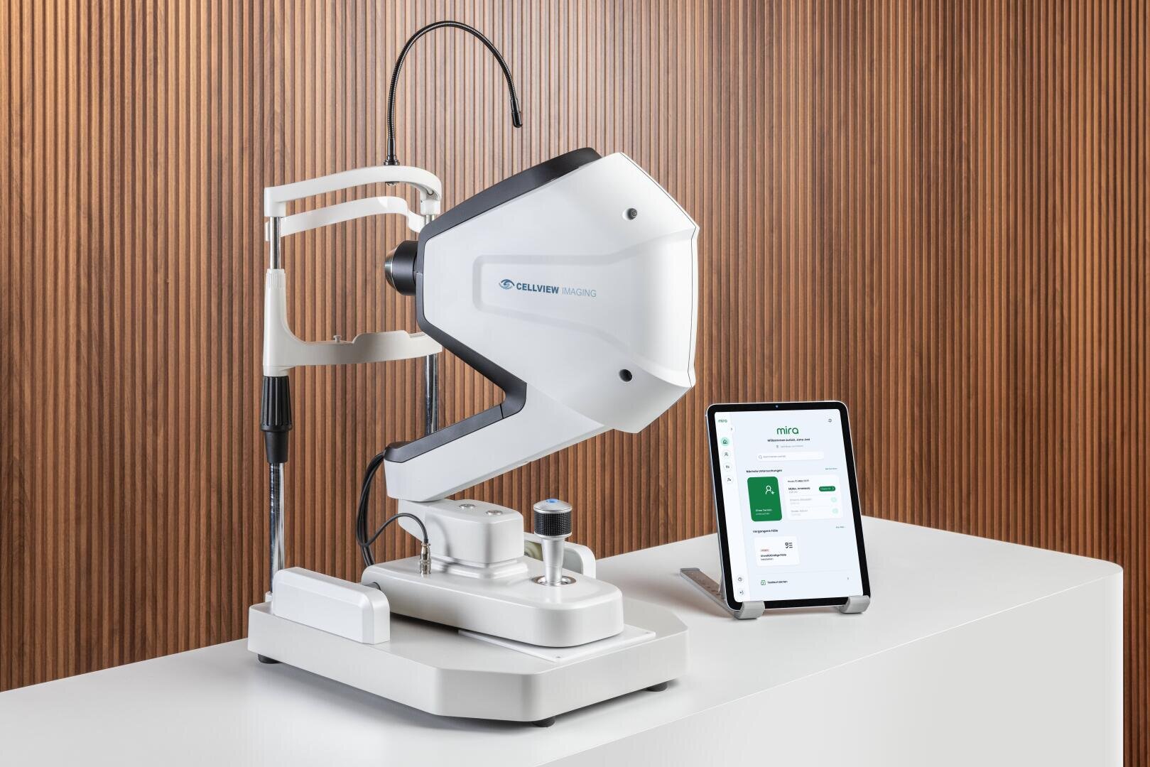 Ein modernes ophthalmologisches Bildgebungsgerät steht auf einem weißen Tresen neben einem Tablet, das eine medizinische App-Schnittstelle anzeigt, mit einer Holztafelwand im Hintergrund.