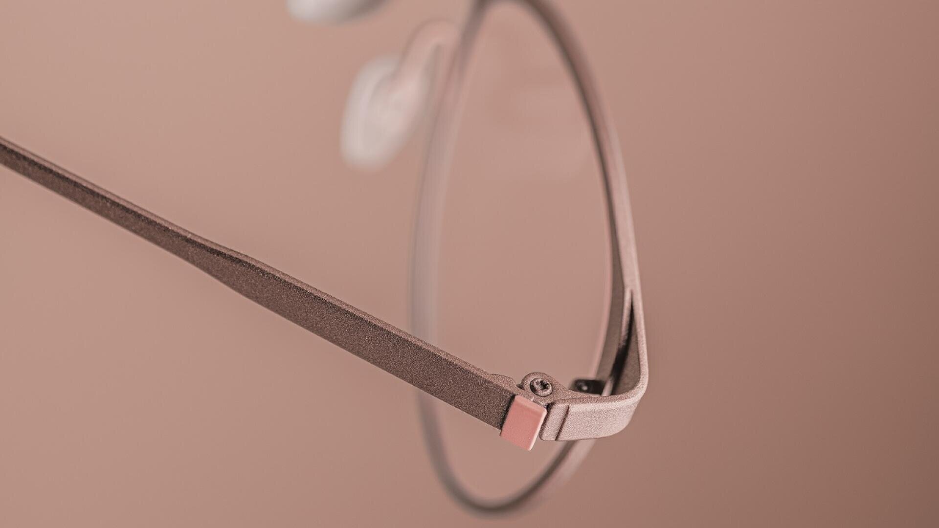 Nahaufnahme einer Brille mit einem dünnen, mattrosa-braunen Rahmen vor einem passenden zartrosa Hintergrund. Nur ein Teil des Glases und der Bügel der Brille sind deutlich zu sehen.
