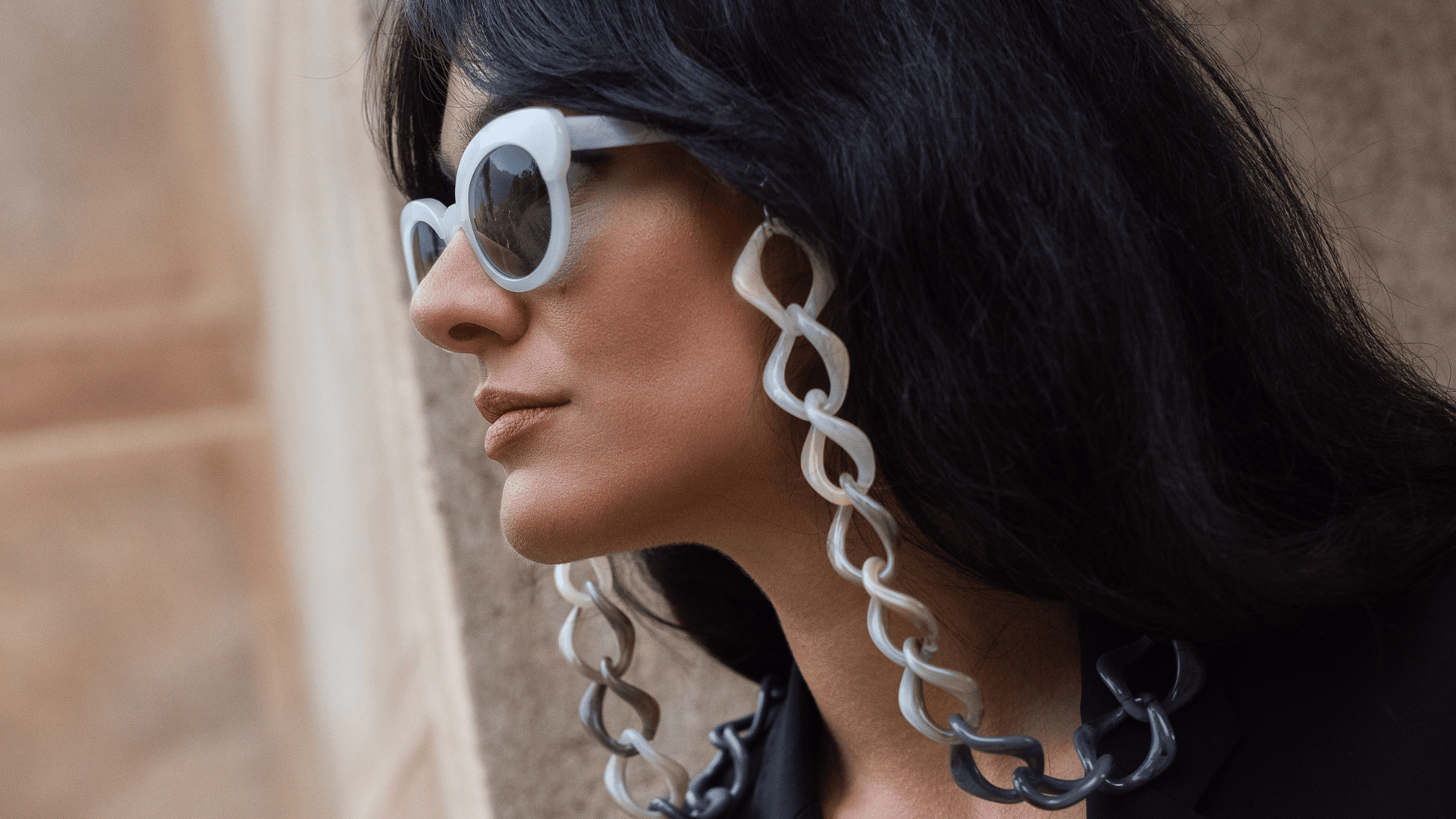 Eine Frau mit langem, dunklem Haar trägt eine übergroße weiße Sonnenbrille mit dicken, klobigen Kettenbefestigungen und ein schwarzes Outfit und steht vor einer Wand in neutraler Farbe.