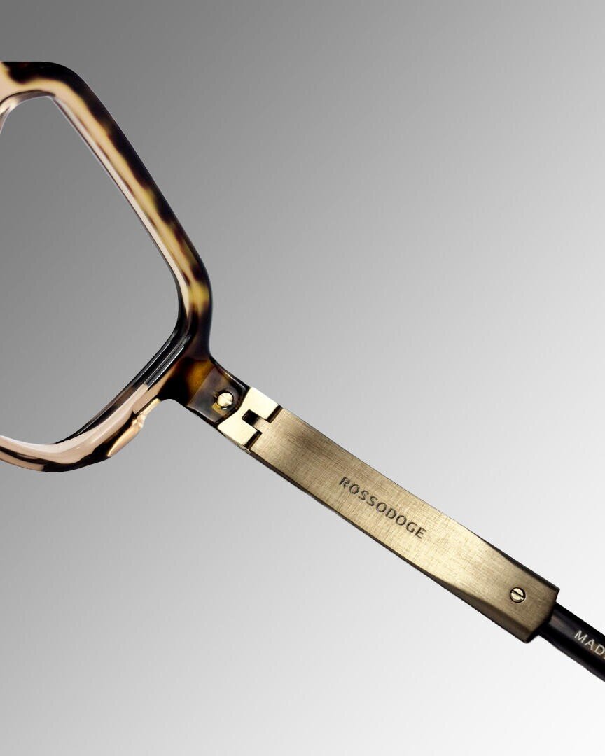 Close-up of tortoiseshell spectacles with a golden metal frame engraved with the brand name ROSSODODGE against a gray background with a gradient.