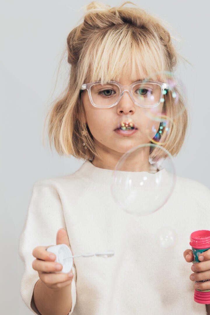 Ein kleines Kind mit blonden Haaren und einer klaren Brille macht Seifenblasen. Es trägt einen hellen Pullover, hält einen Seifenblasenstab und einen rosa Behälter in der Hand und konzentriert sich konzentriert auf die schwebenden Blasen.