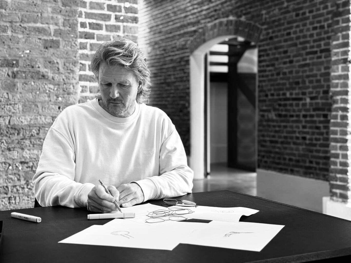 Ein Mann in einem weißen Sweatshirt sitzt an einem Tisch und zeichnet mit einem Stift auf Papier. Mehrere Blätter mit Skizzen und eine Brille liegen auf dem Tisch. Er befindet sich in einem Raum mit freiliegenden Ziegelwänden und gewölbten Türöffnungen.