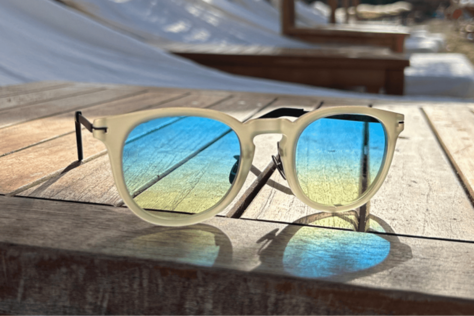 Eine weiß umrandete Sonnenbrille mit blau-gelben Verlaufsgläsern liegt auf einem Holztisch und reflektiert das Sonnenlicht. Im Hintergrund sind unscharfe Strandkörbe und weiße Stoffschirme zu sehen.