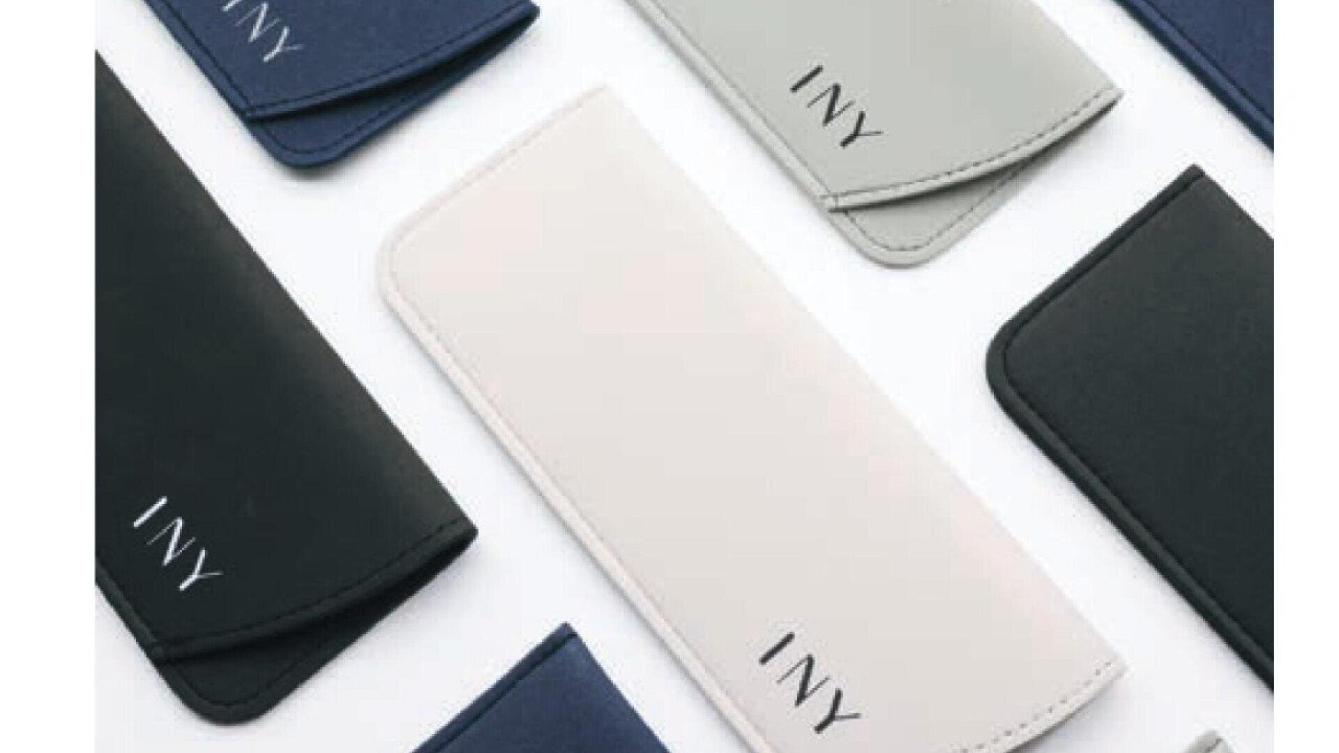 Several sleek, minimalist eyeglass cases in various colors (black, navy, gray, and white) with the letters INY printed on them, arranged diagonally on a white surface.