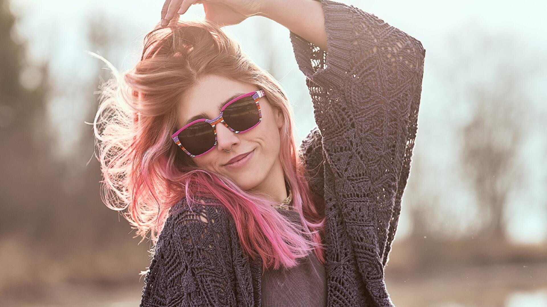 Eine Person mit langen rosafarbenen Haaren und Sonnenbrille lächelt, während sie ihr Haar hochhält. Sie trägt einen strukturierten, dunklen Strickpullover und steht im Freien im sanften Sonnenlicht.
