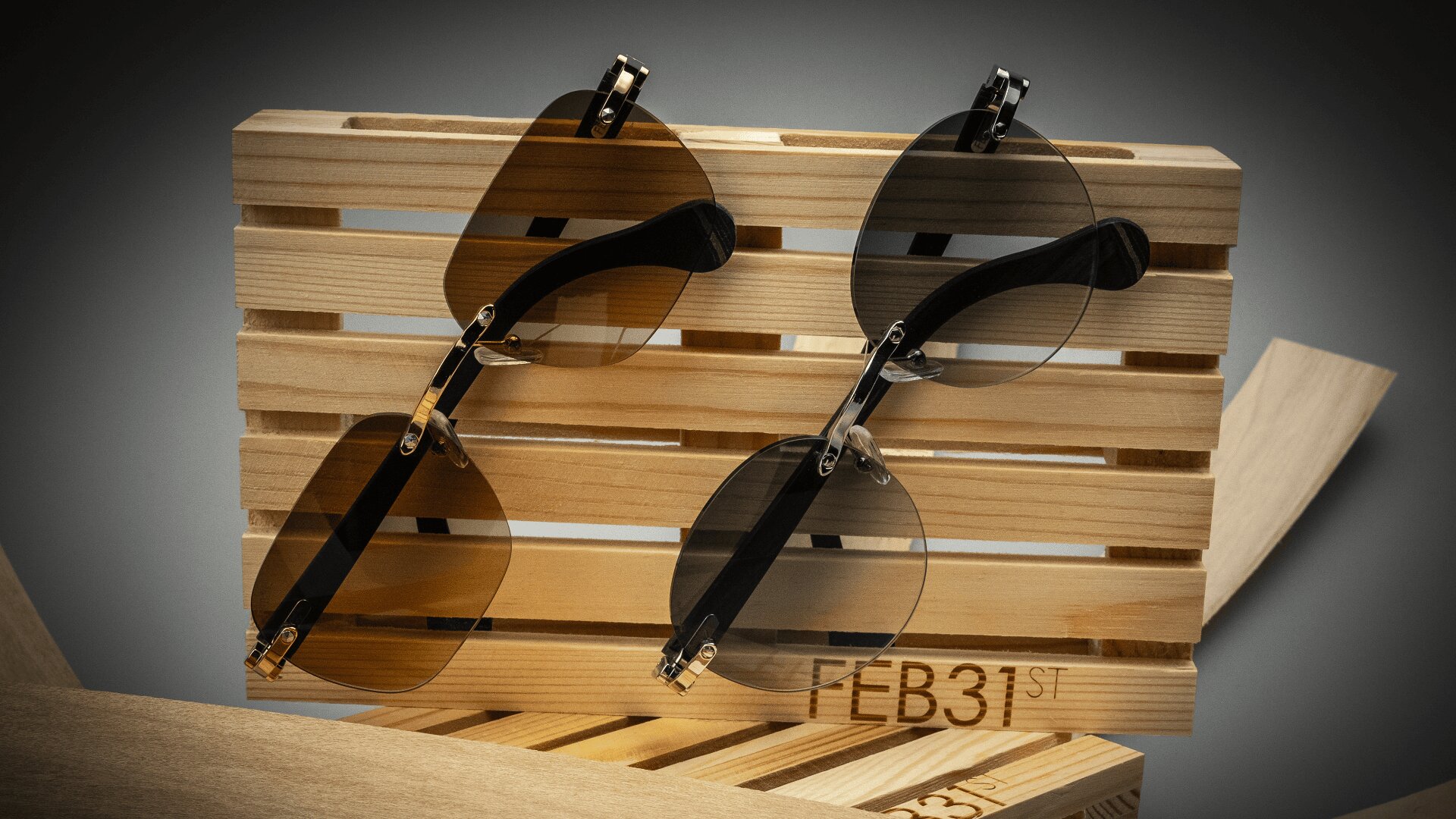 Zwei randlose Sonnenbrillen, eine mit braunen und eine mit grauen Gläsern, stehen auf einem Holzständer mit der Gravur FEB31st. Der Hintergrund ist grau und besteht aus Holzbrettern.