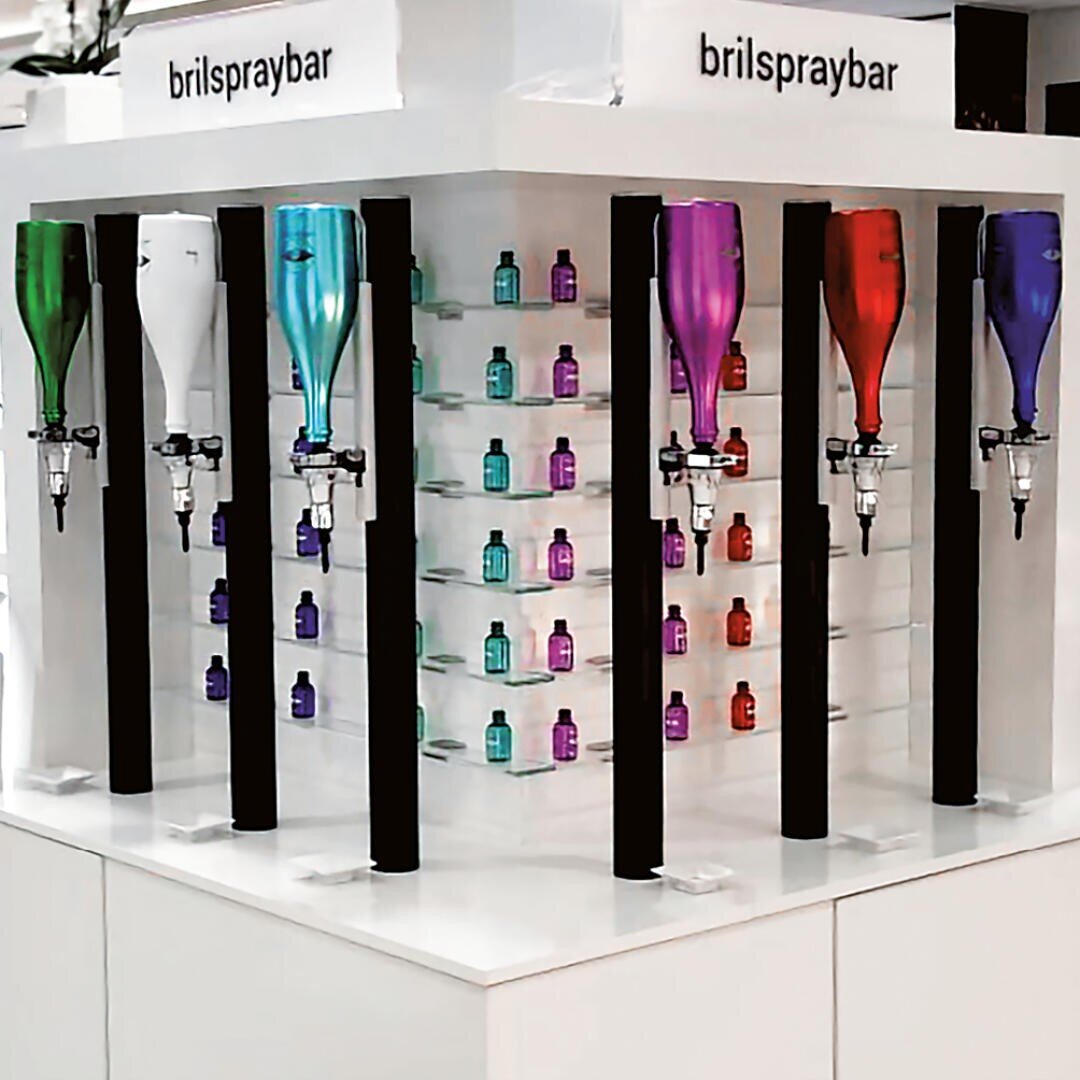 An einer Bartheke stehen sechs große, bunte Flaschen mit Ausgießern zum Ausschenken von Flüssigkeiten. Dahinter reihen sich in Regalen kleine, bunte Flaschen in Blau, Lila, Pink und Grün. Auf den Schildern steht „Brilspraybar“.